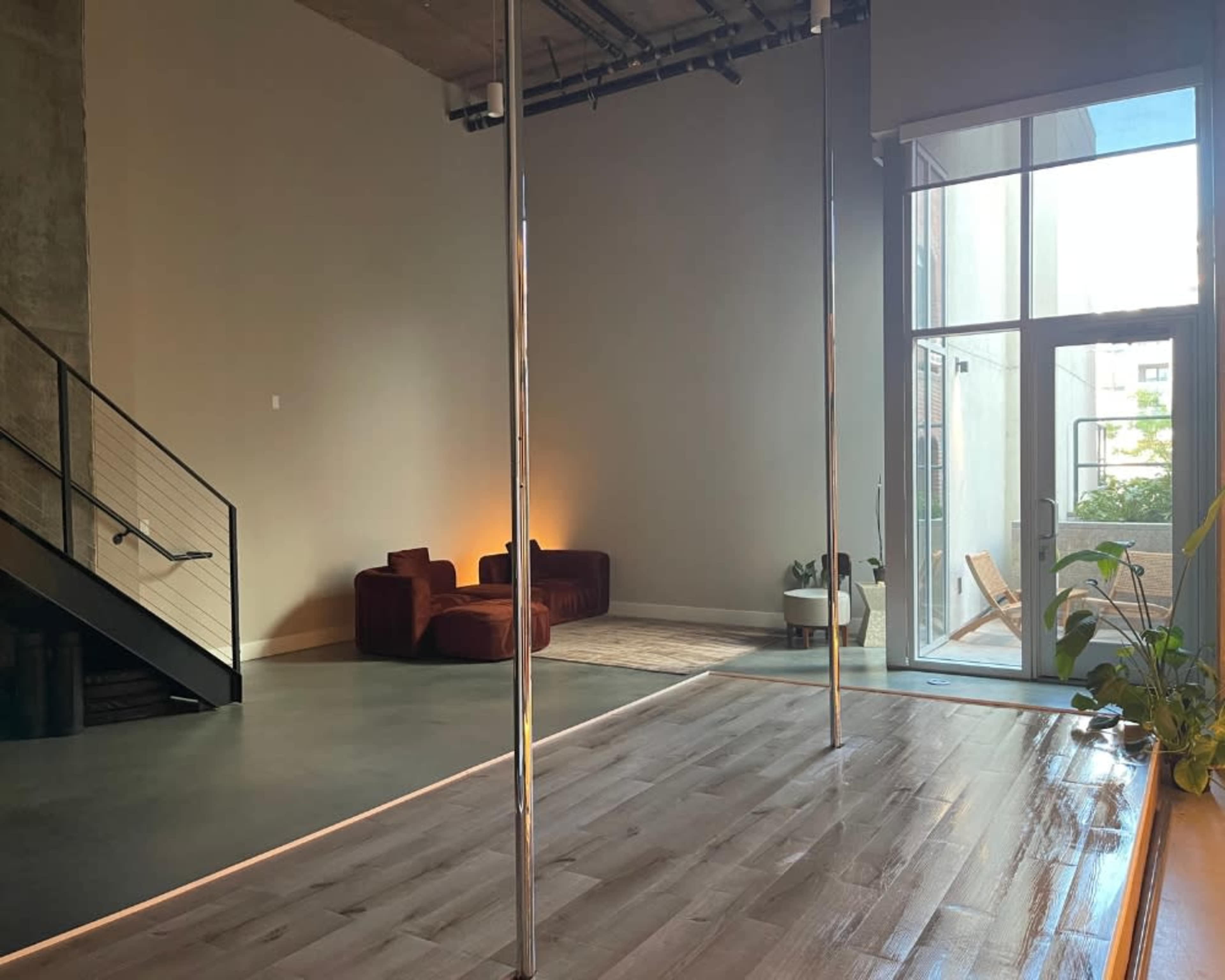 The image shows a spacious room with hardwood flooring, a pole in the center, a staircase on the left, and a seating area with red couches in the background.