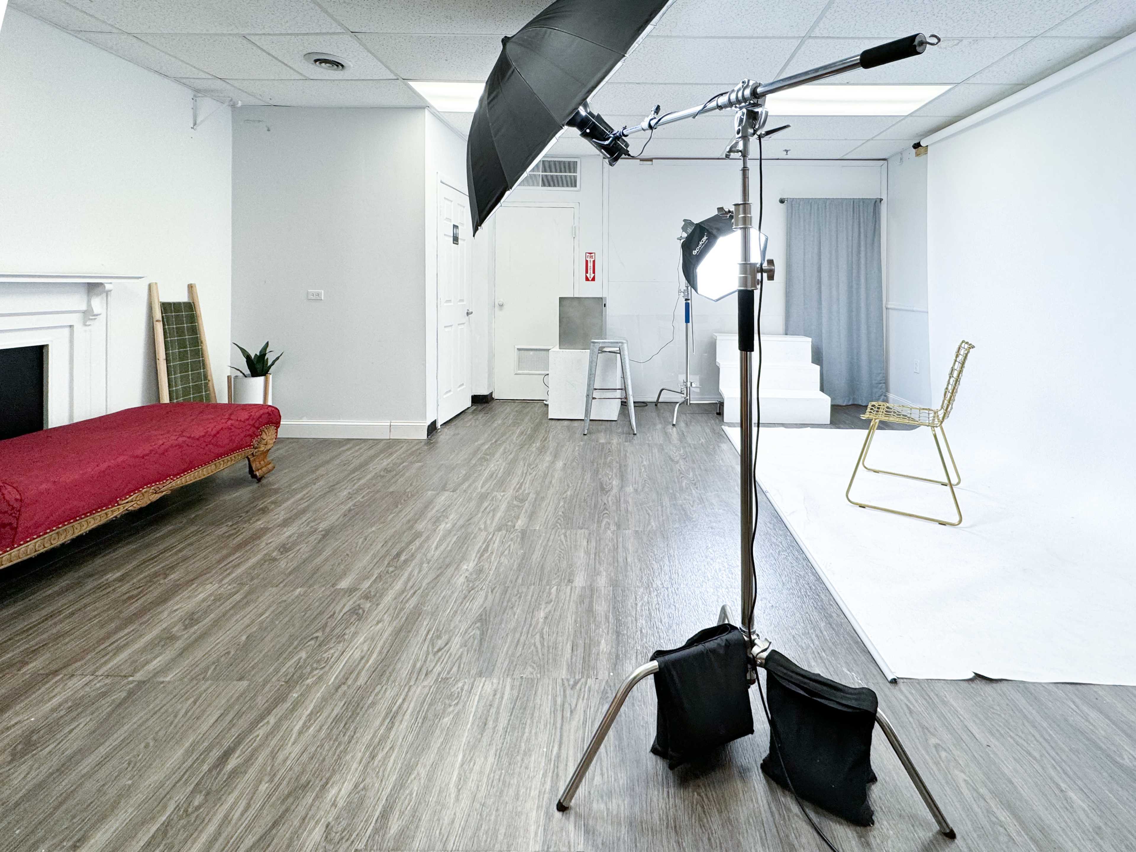 A photography studio with a wooden floor, a red couch, a gold chair, and a large umbrella light setup.