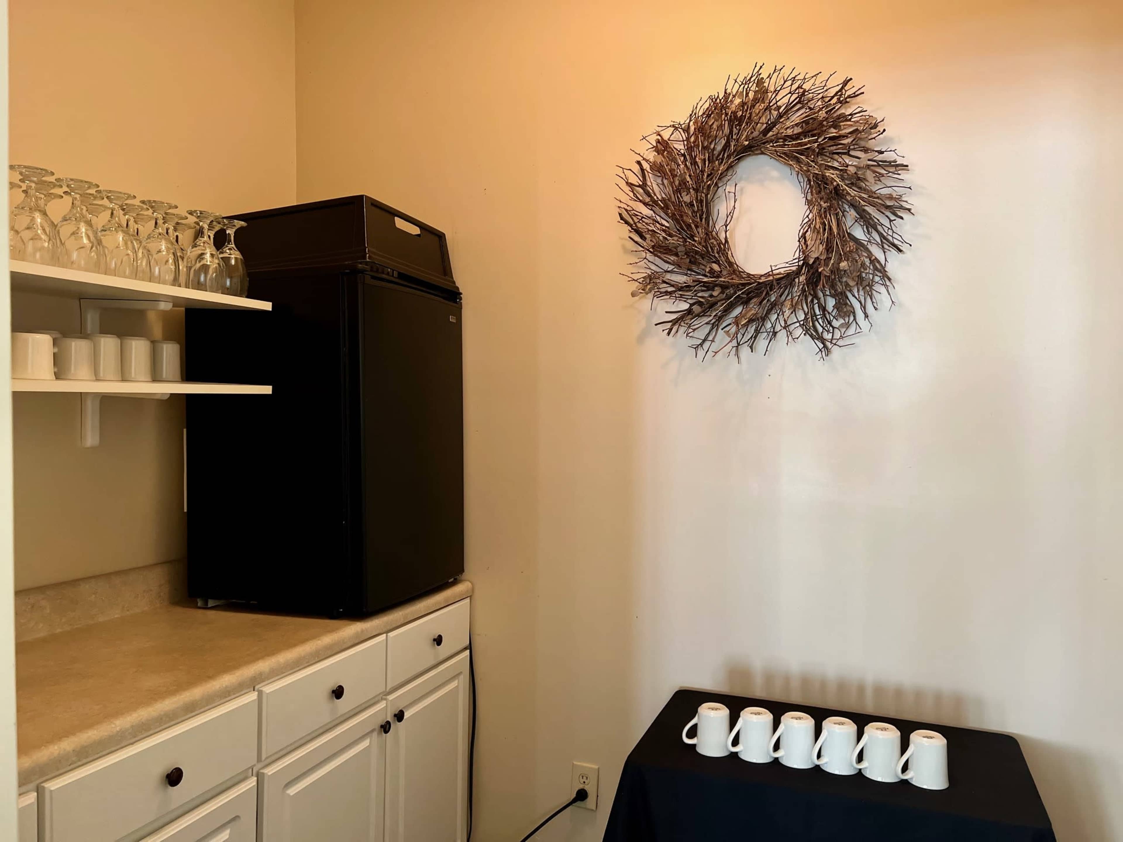 A small room features a black refrigerator, a countertop with glassware, and a wreath mounted on the wall above a table displaying several white cups.