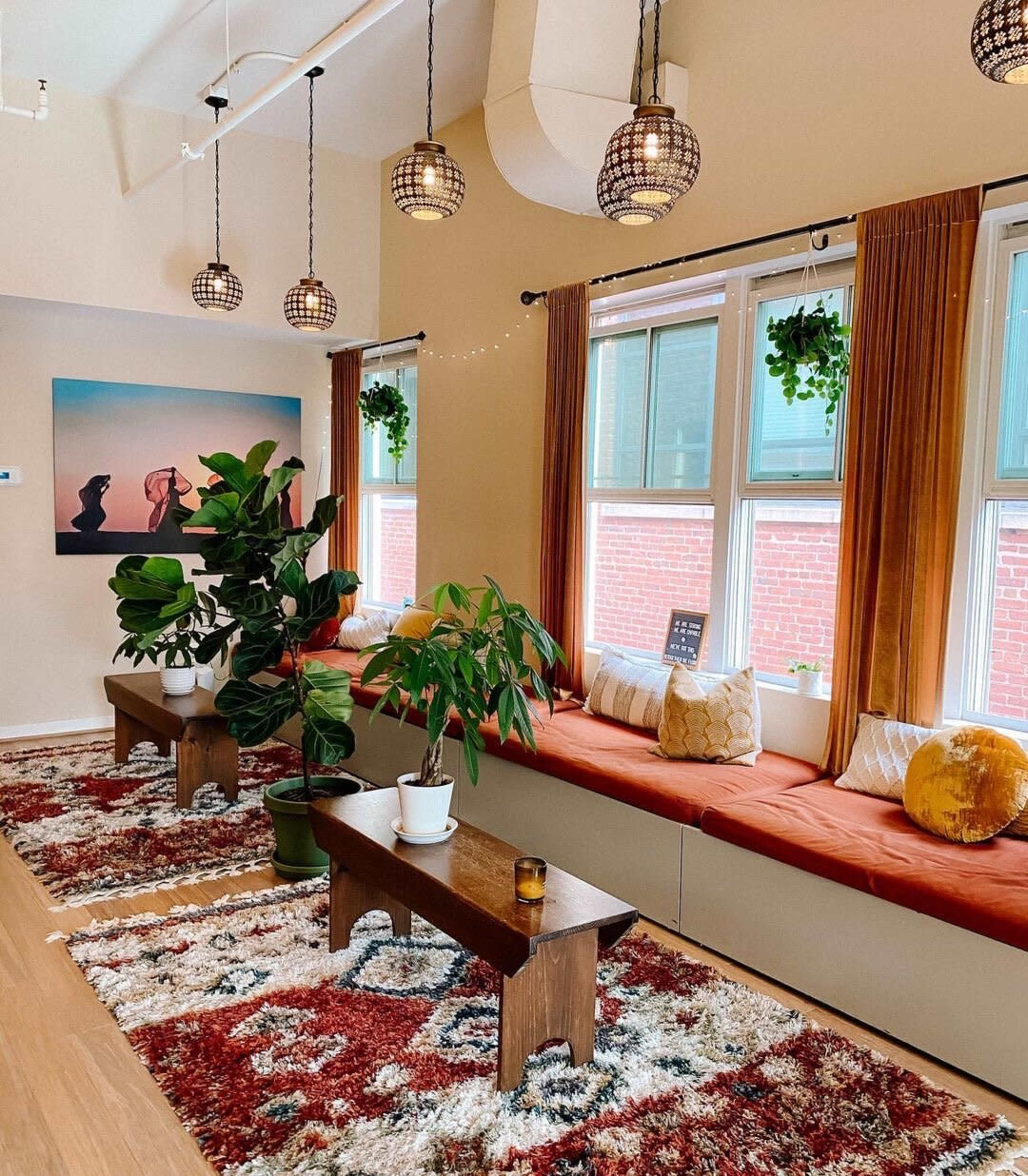 A spacious, well-lit room with two windows, wooden benches adorned with cushions, indoor plants, and decorative rugs.