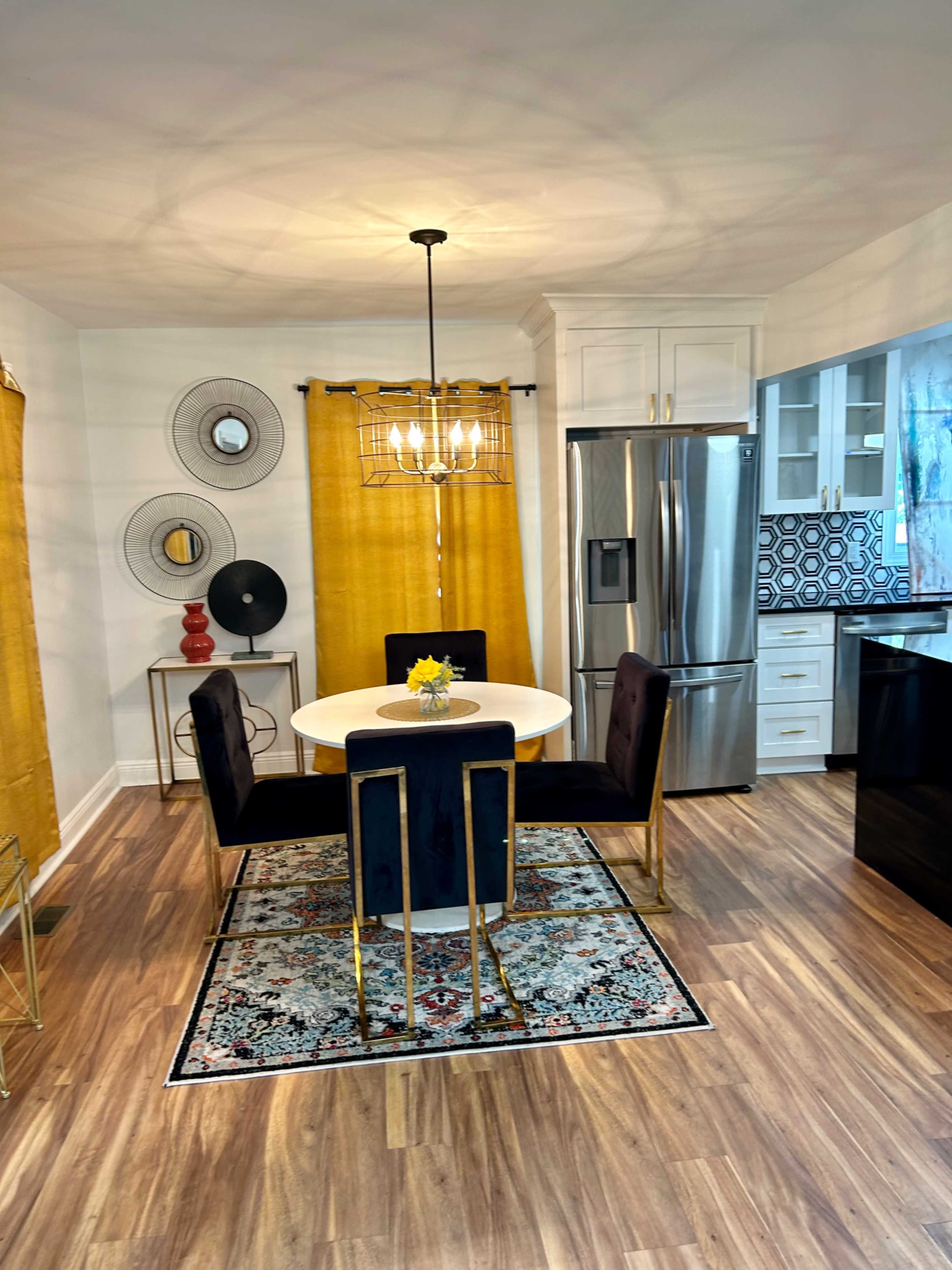 A modern kitchen with a circular dining table surrounded by four black velvet chairs, yellow curtains, and a decorative rug.
