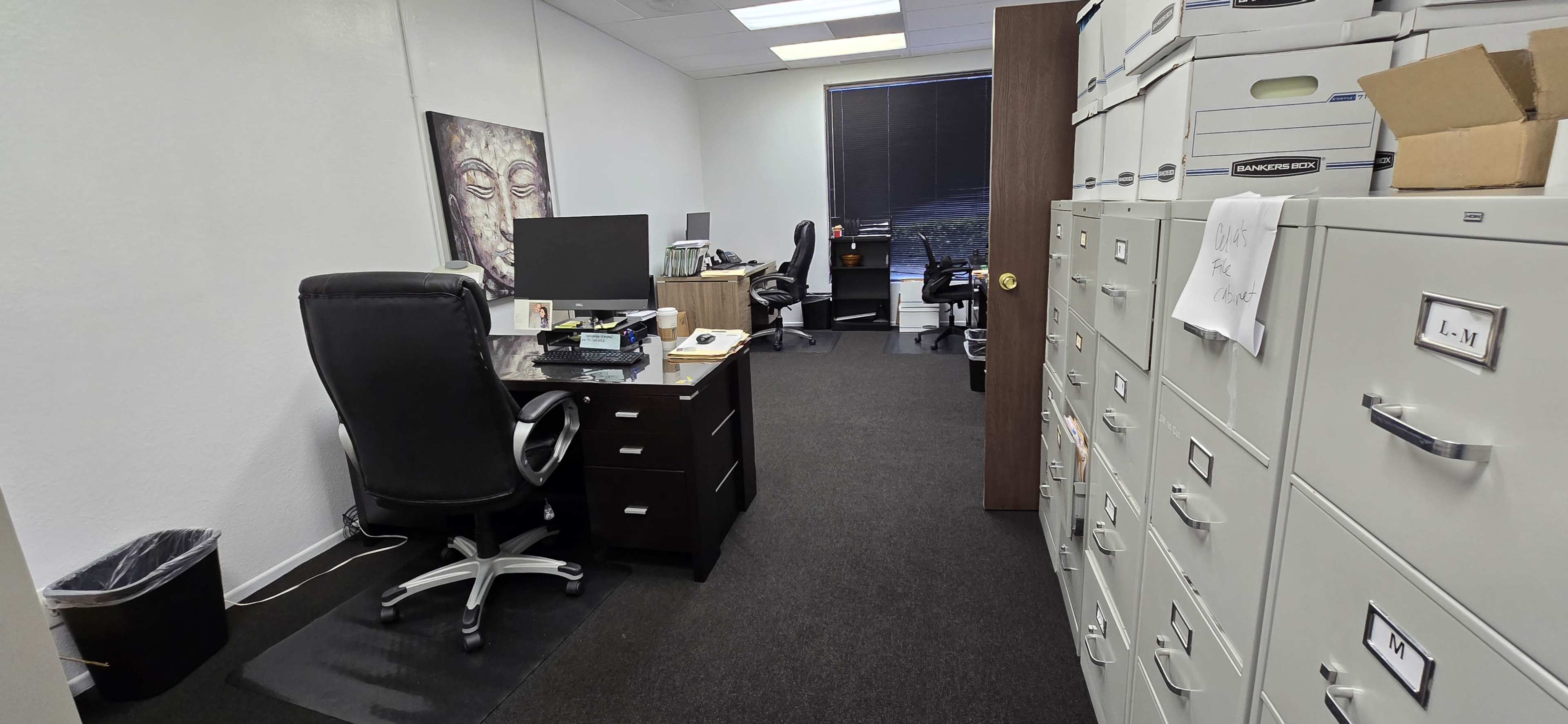An office space featuring filing cabinets, a desk with a computer, and two chairs visible in the background.