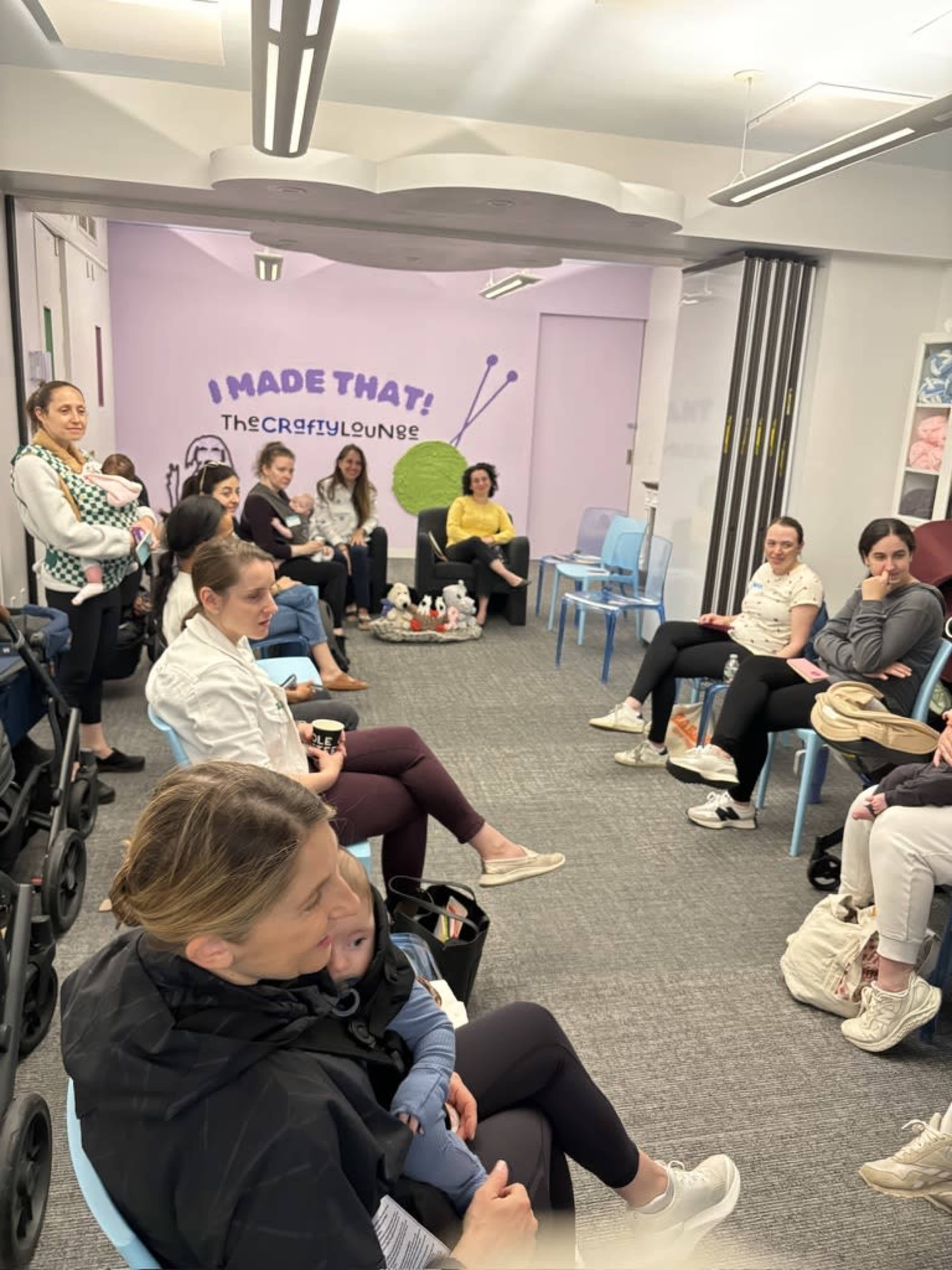 A group of people is seated in a brightly lit room with a purple wall that features a crafting-related mural.