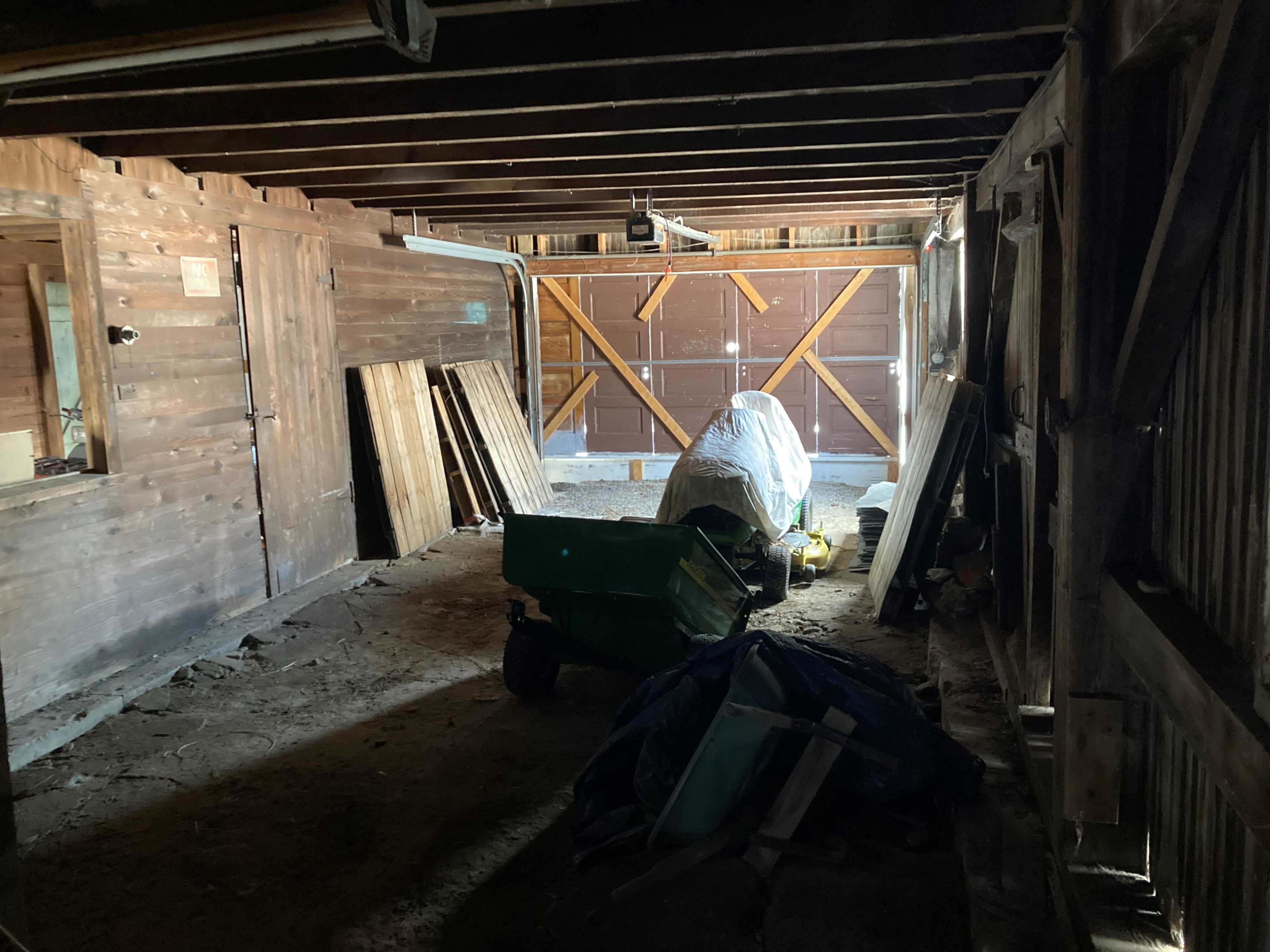 The interior of a dimly lit barn features wooden beams, storage materials against the walls, and a covered vehicle at the center.