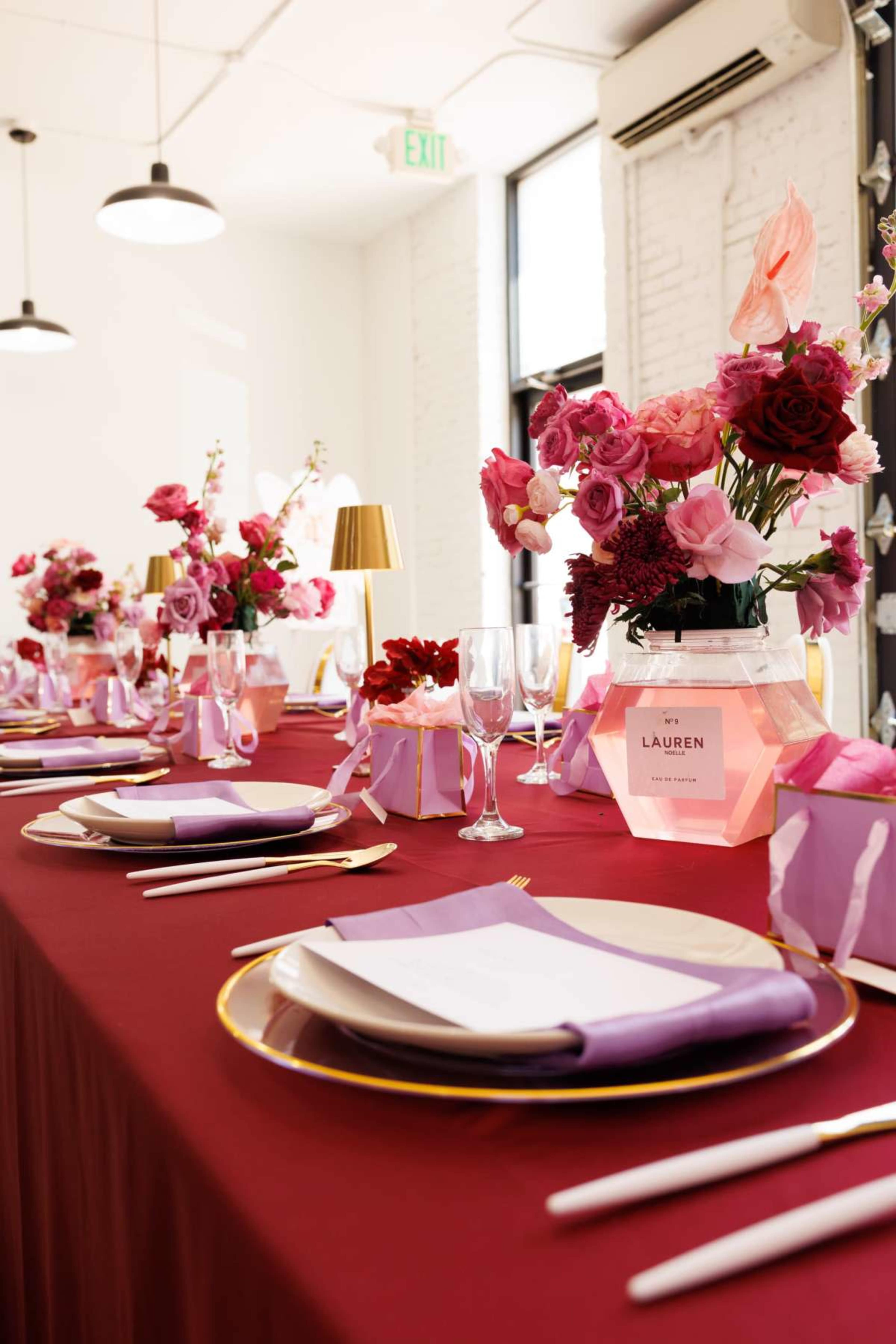 A long table is set with elegant place settings, featuring gold-rimmed plates and floral centerpieces in shades of pink and purple.