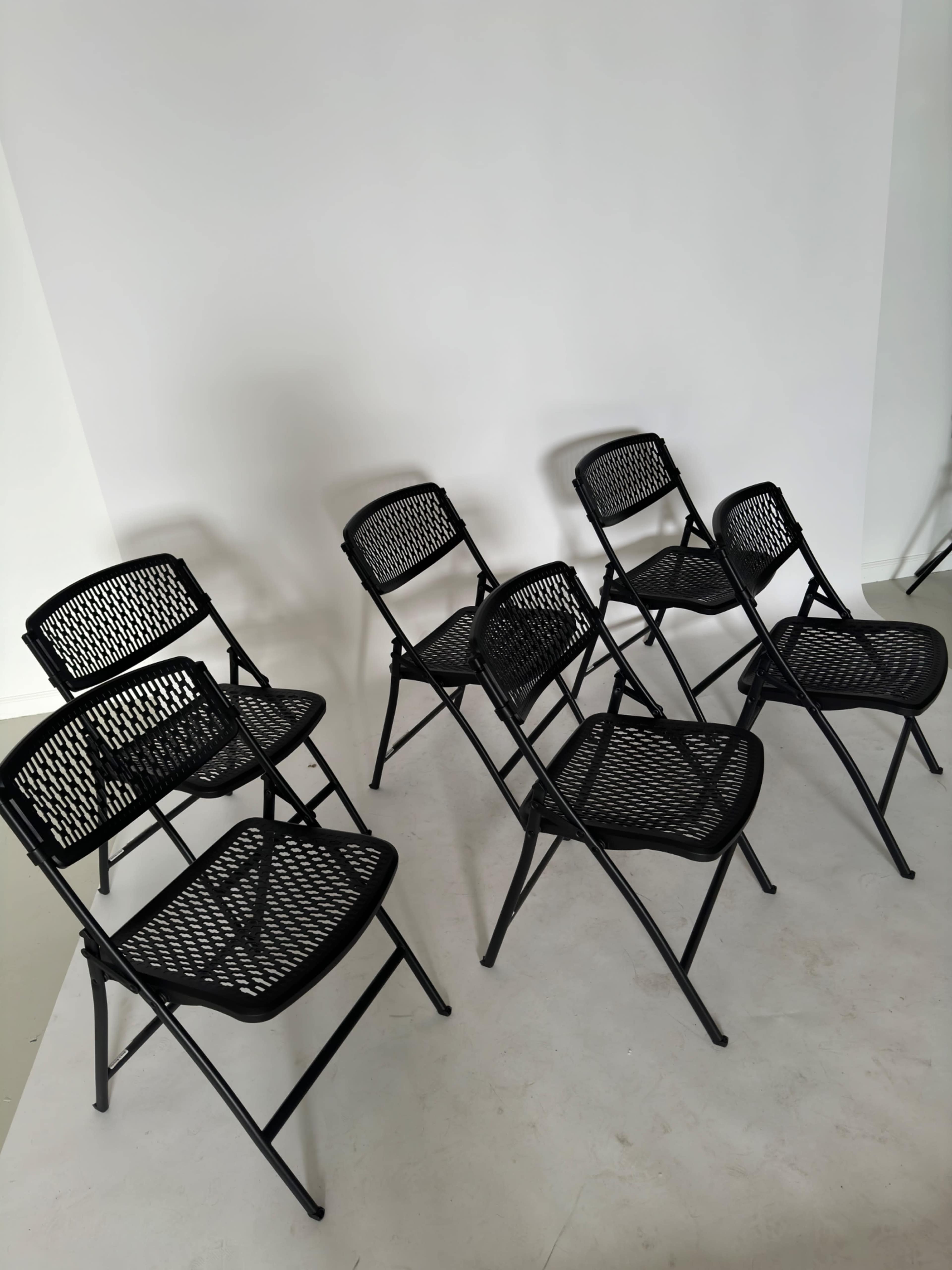 Six black folding chairs arranged against a plain white wall.