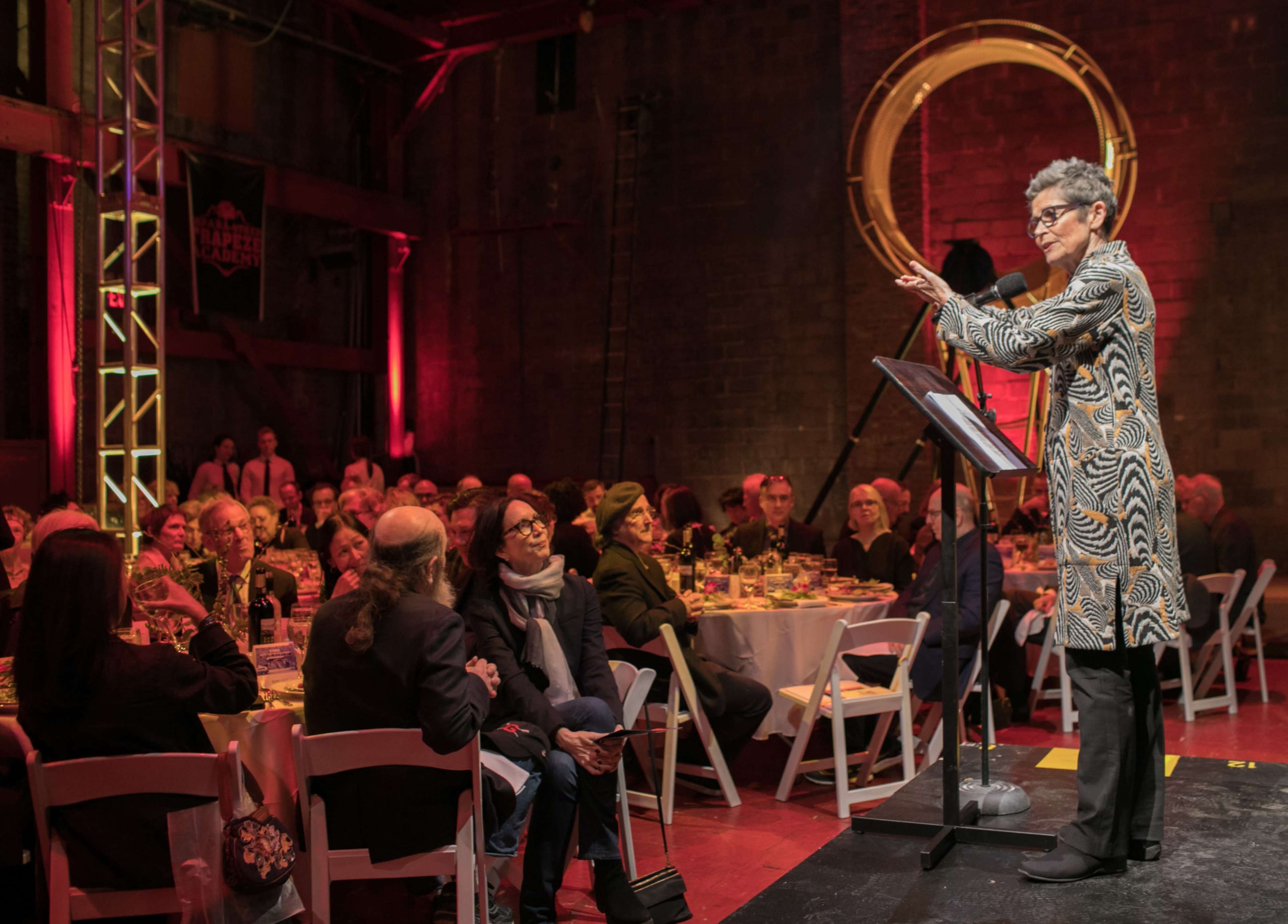 A speaker stands at a podium addressing an audience seated at tables during an event in an industrial space.