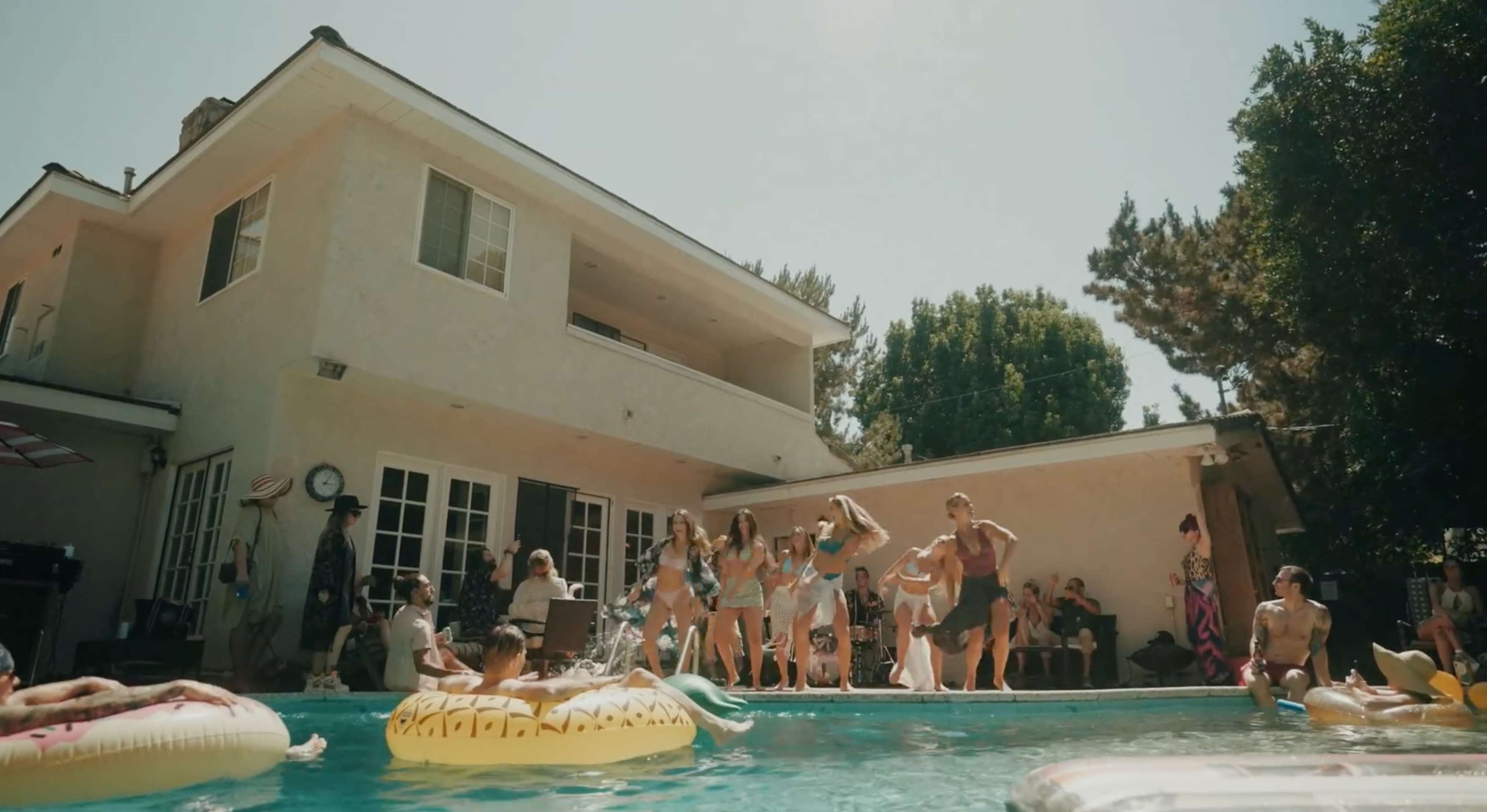 A group of people gather around a swimming pool at a house party on a sunny day, with some swimming and others socializing on the pool deck.