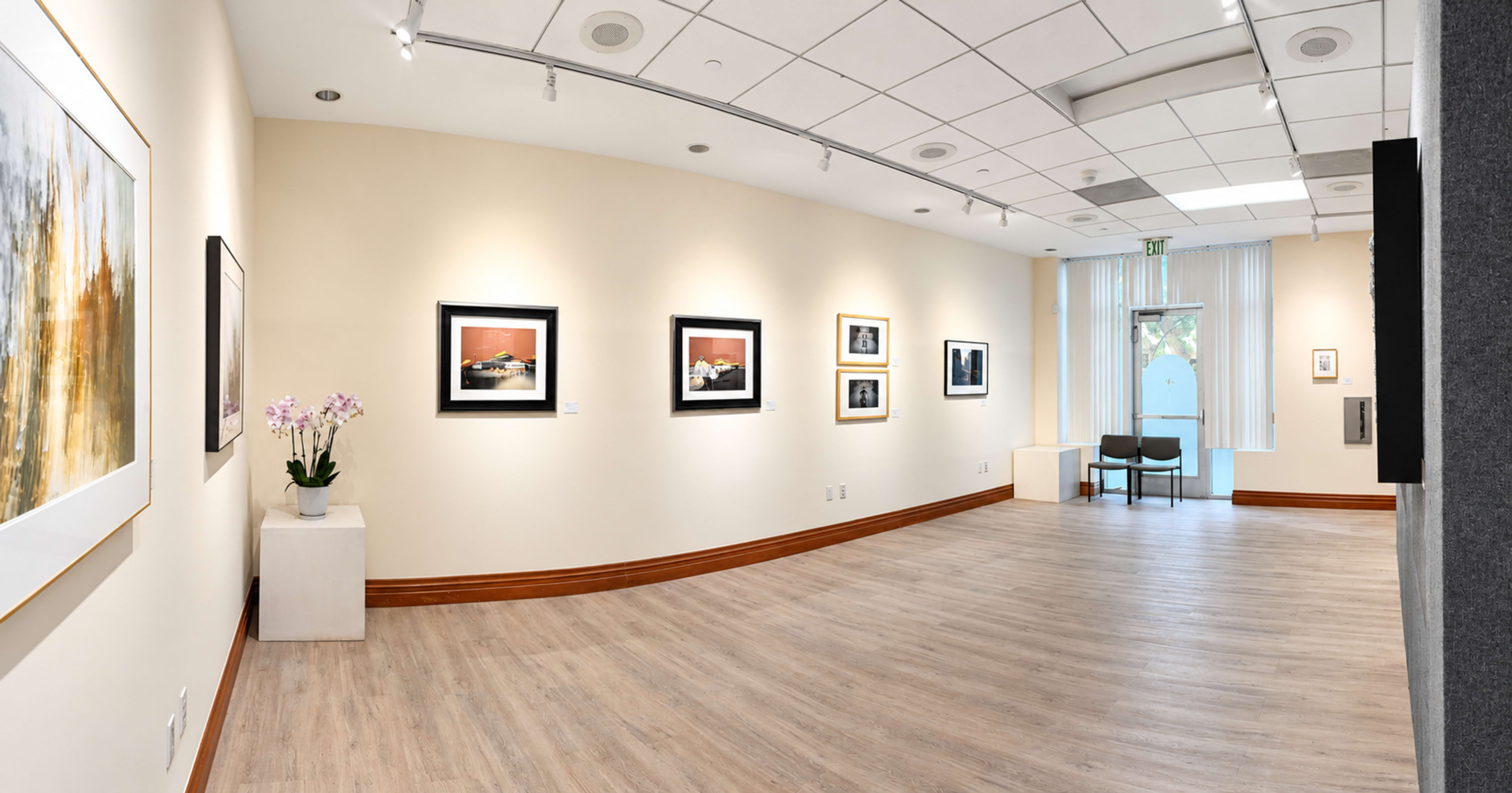 The image depicts a well-lit art gallery featuring several framed photographs hanging on the walls and a small seating area in the corner.