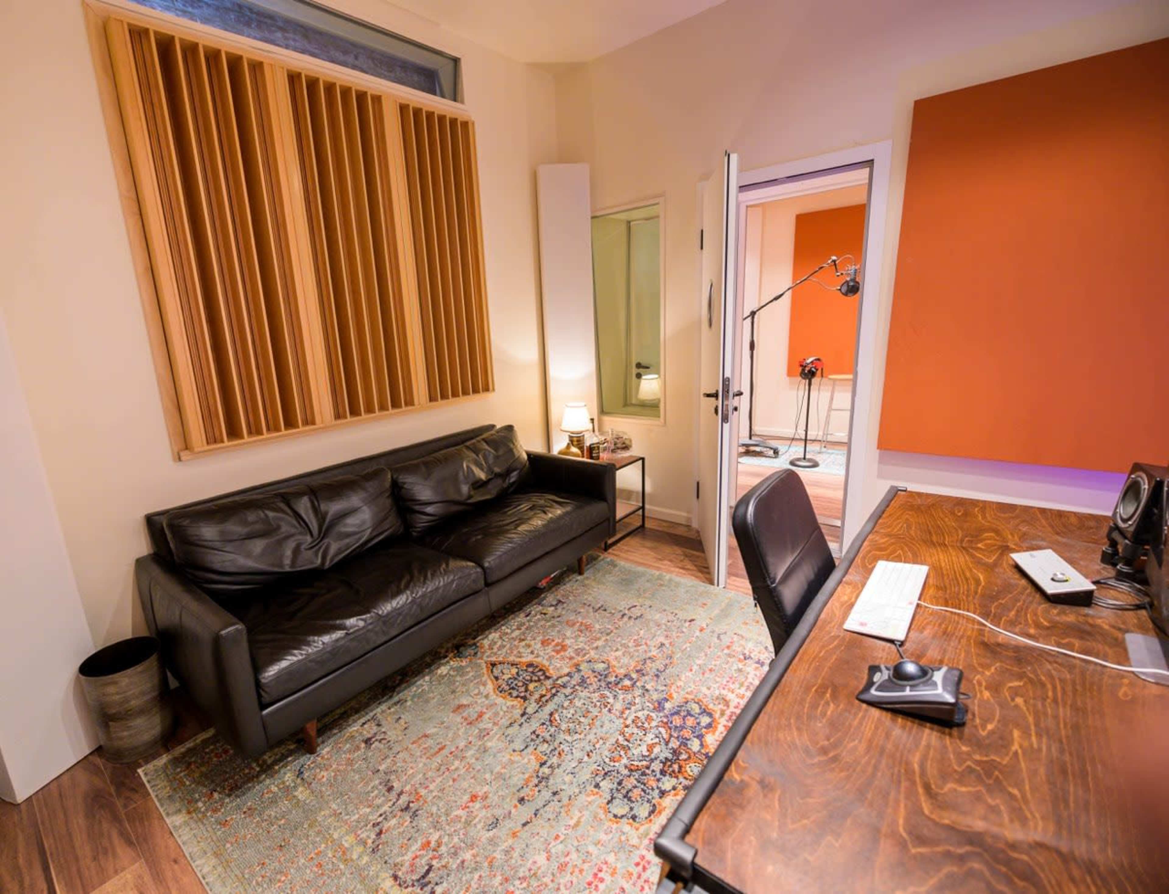 The image shows a recording studio with a black leather sofa, a wooden desk with equipment, and soundproofing panels on the walls.