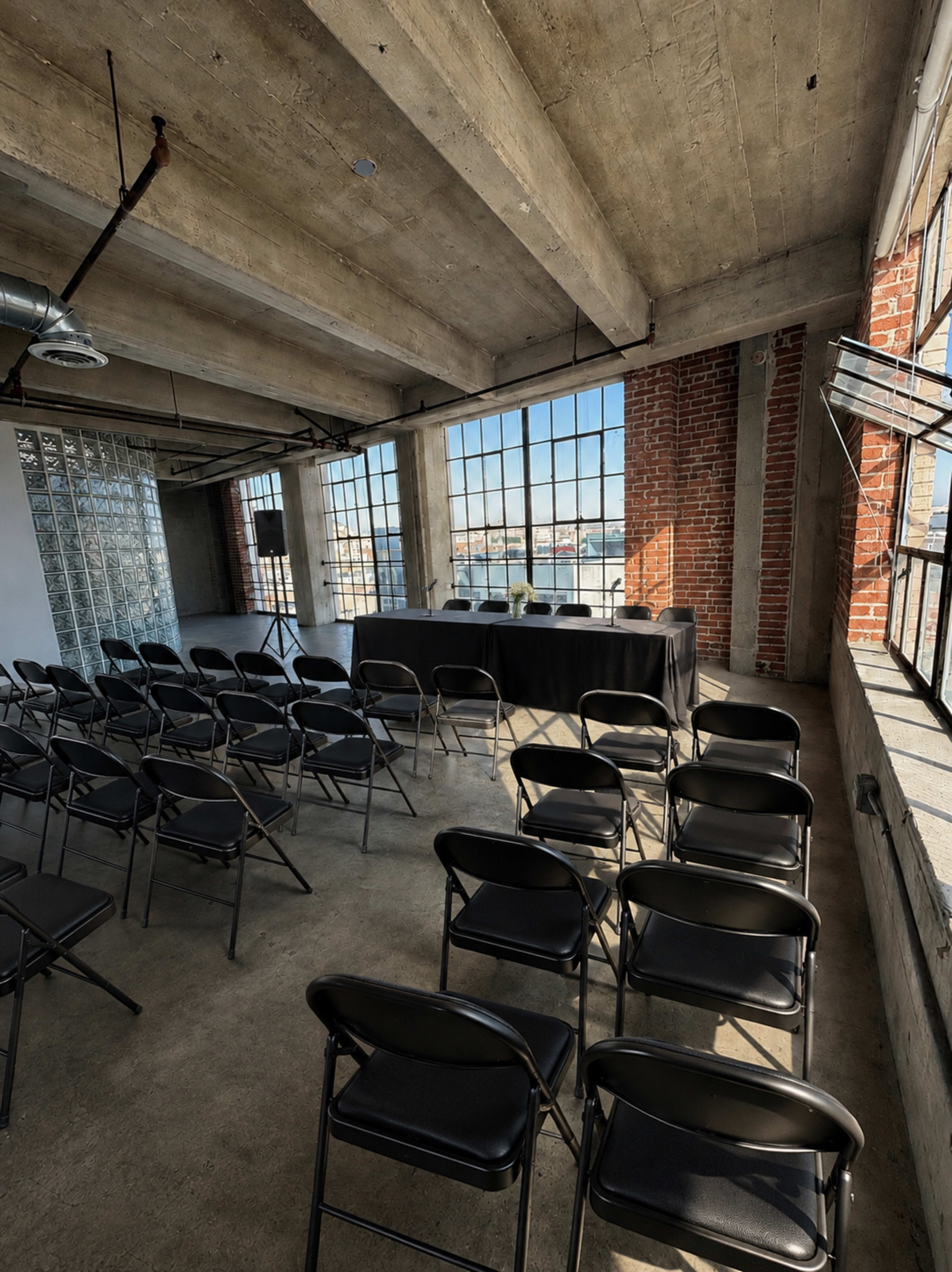 The image shows a spacious room with tall windows, a table set up at the front, and rows of black folding chairs arranged for an event.