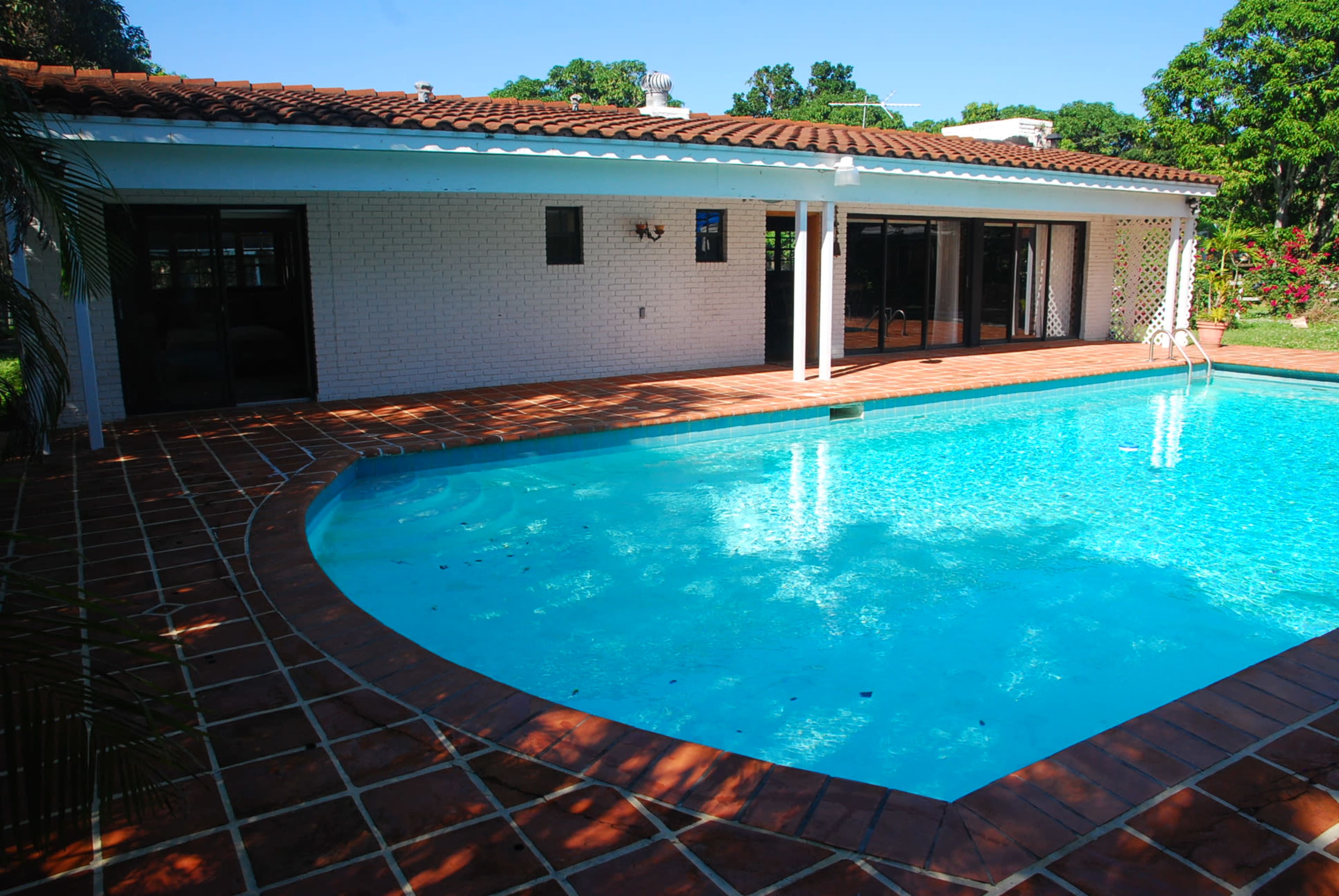 A rectangular swimming pool with clear blue water is situated beside a single-story white house, surrounded by a brick patio and green landscaping.