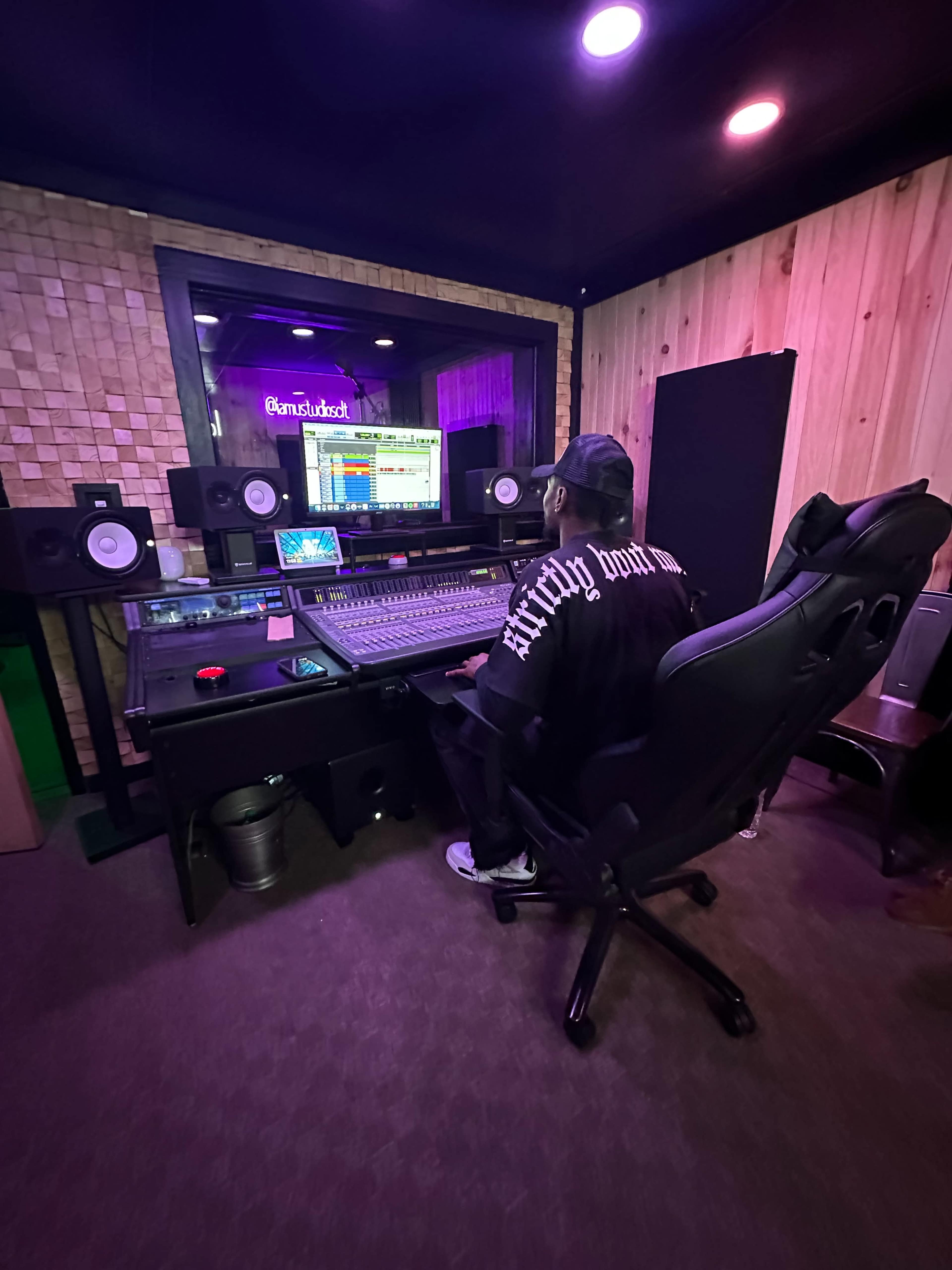 A person sits at a sound mixing desk in a recording studio, surrounded by audio equipment and monitors.