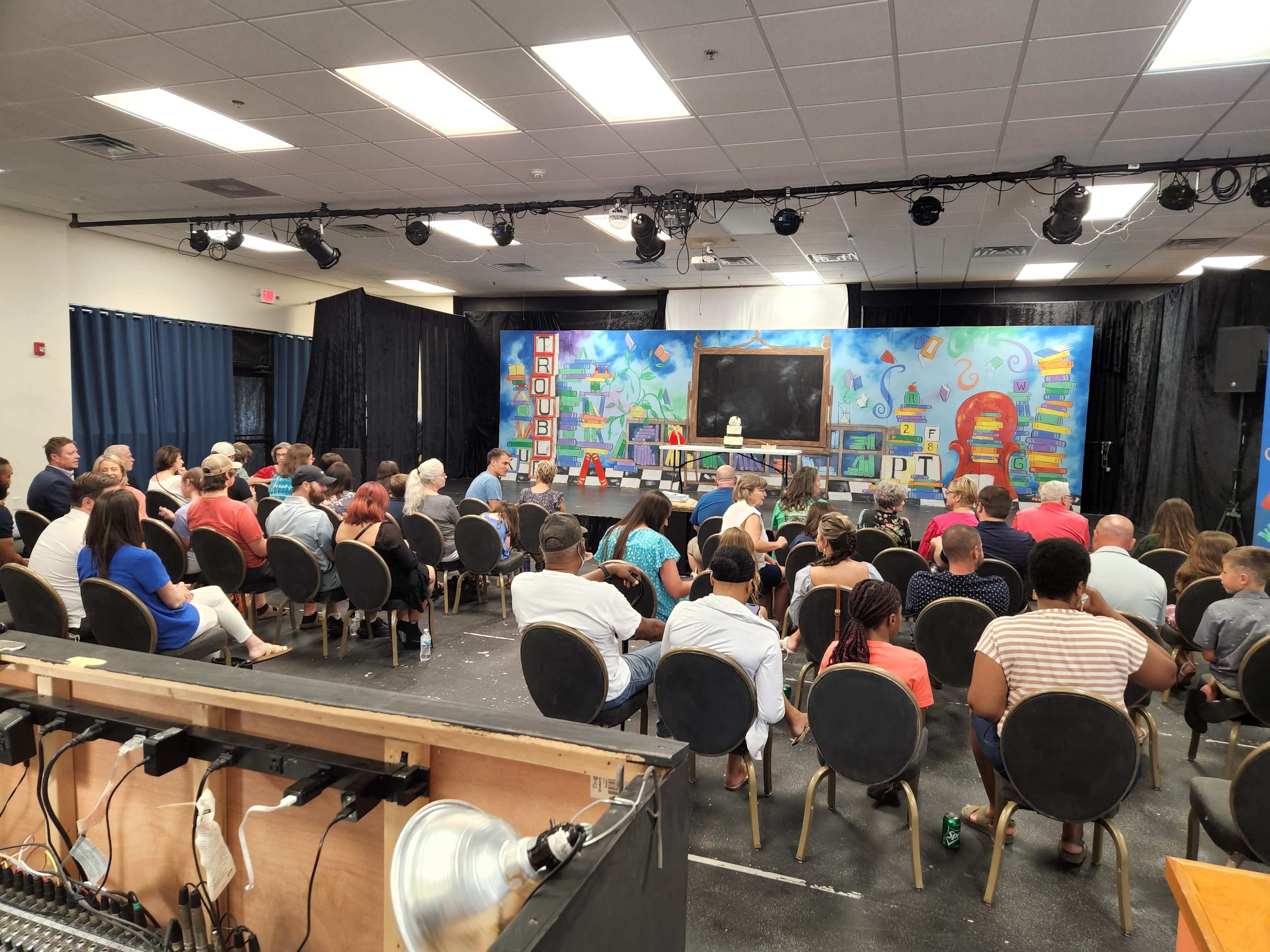 A group of people sits in rows facing a stage adorned with a colorful backdrop featuring various educational elements.