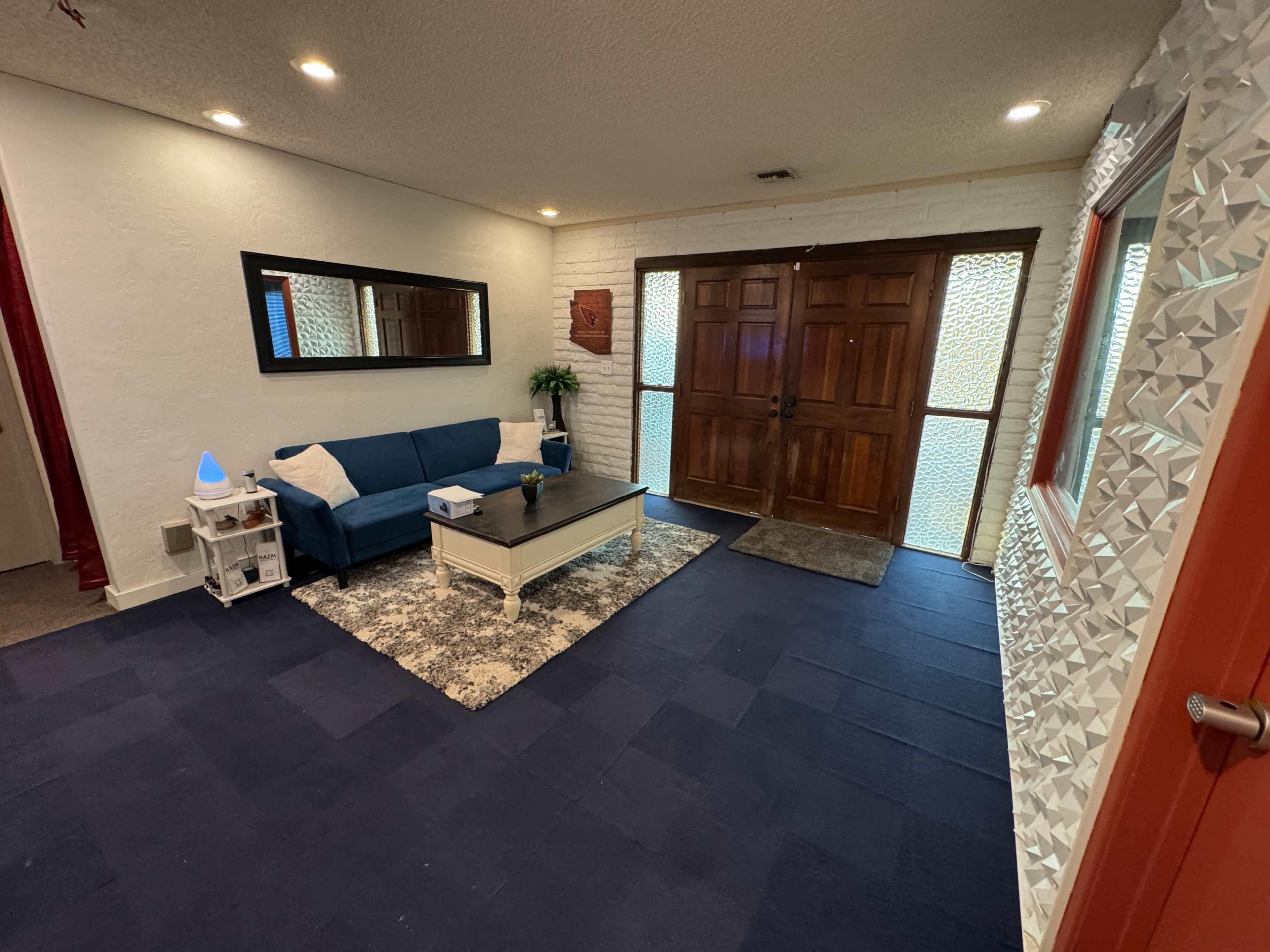 The image shows a spacious entryway with a blue couch, a coffee table, and double wooden doors that open to the entrance.