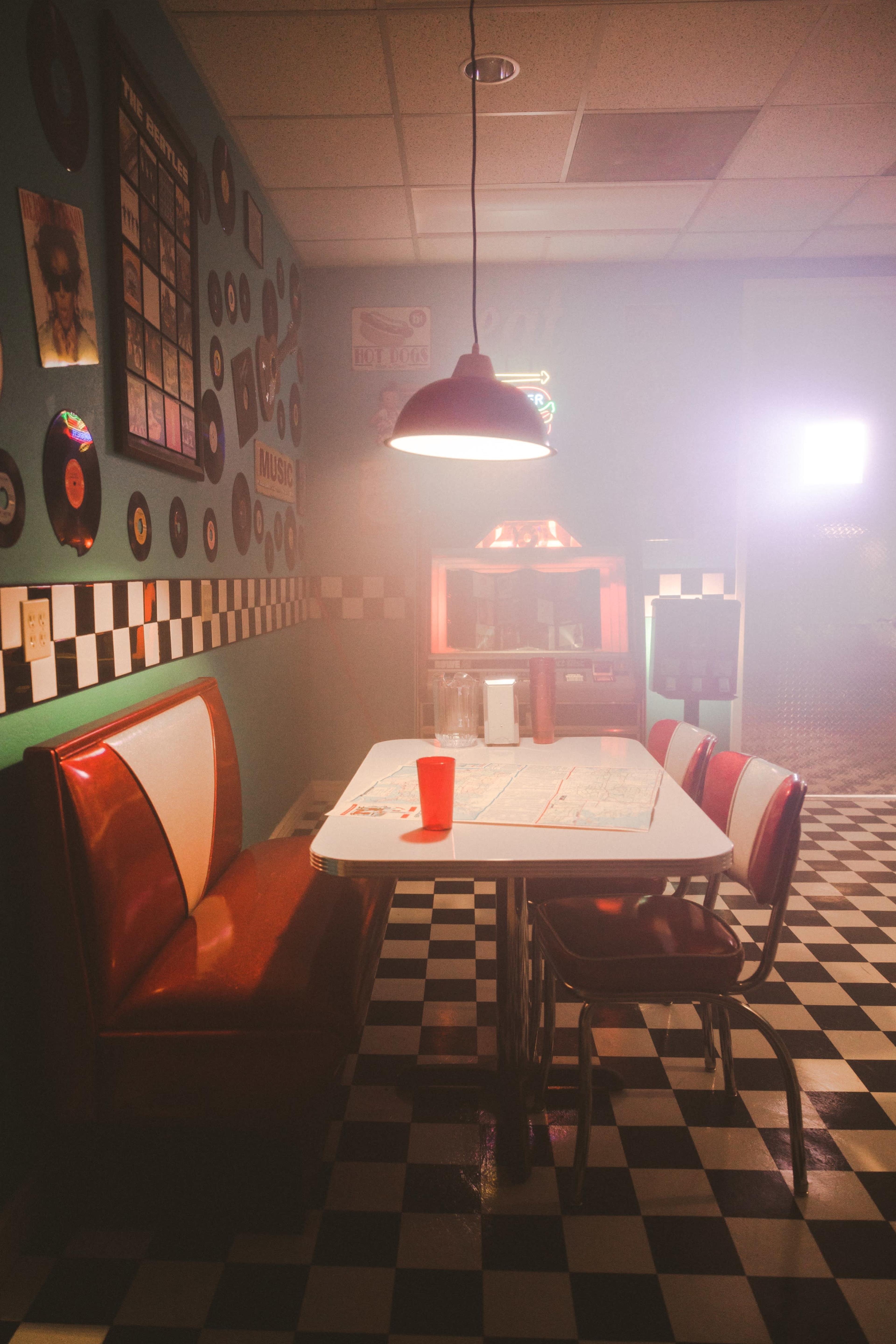 A vintage diner scene features a red booth and a white table set on black-and-white checkered flooring, illuminated by a hanging light, with a red cup on the table.
