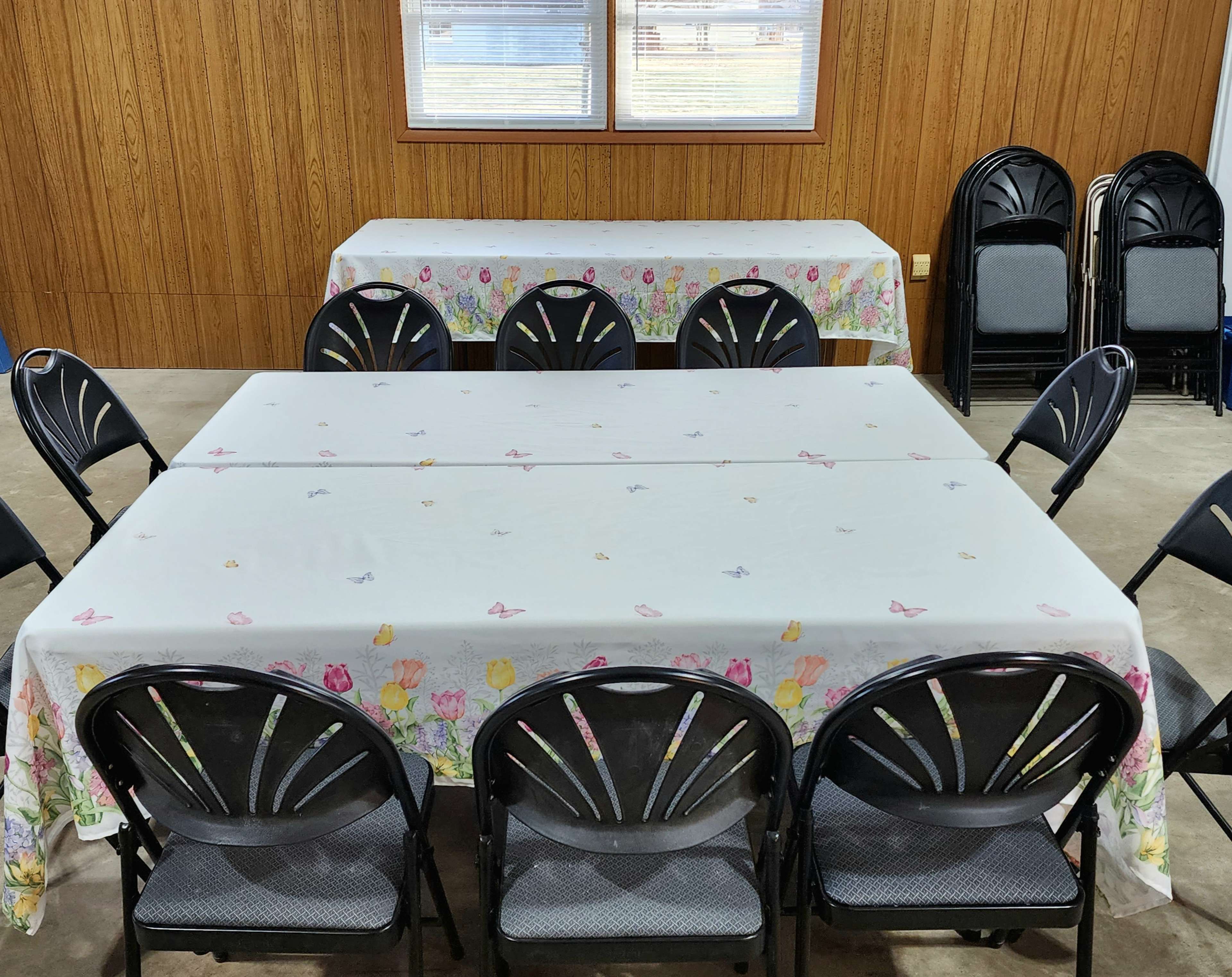 Several tables covered with floral tablecloths are set up in a room with black folding chairs arranged around them.