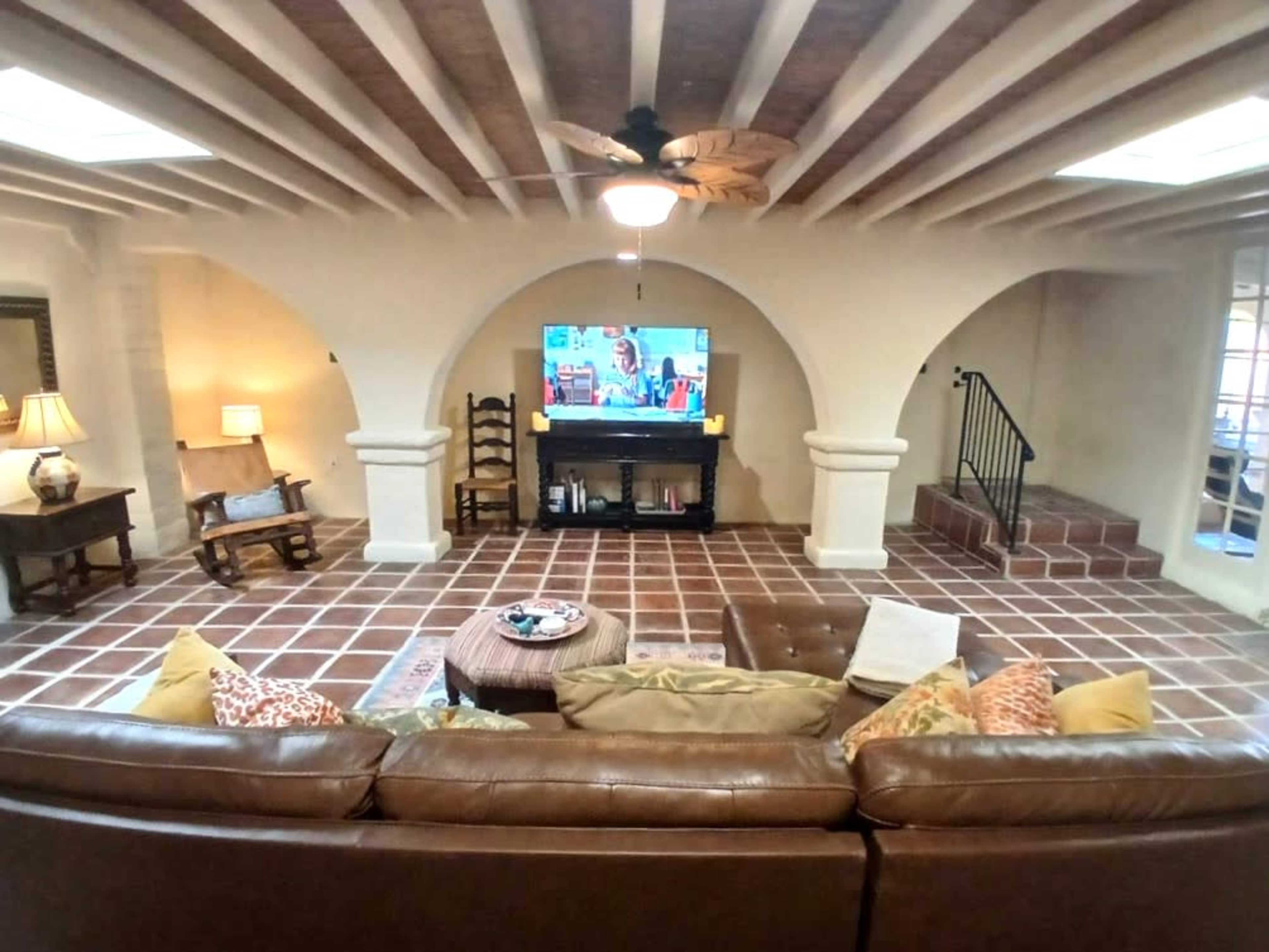 The living room features a leather sofa facing a wall-mounted television, with ceramic tile flooring and arched doorways leading to other spaces.