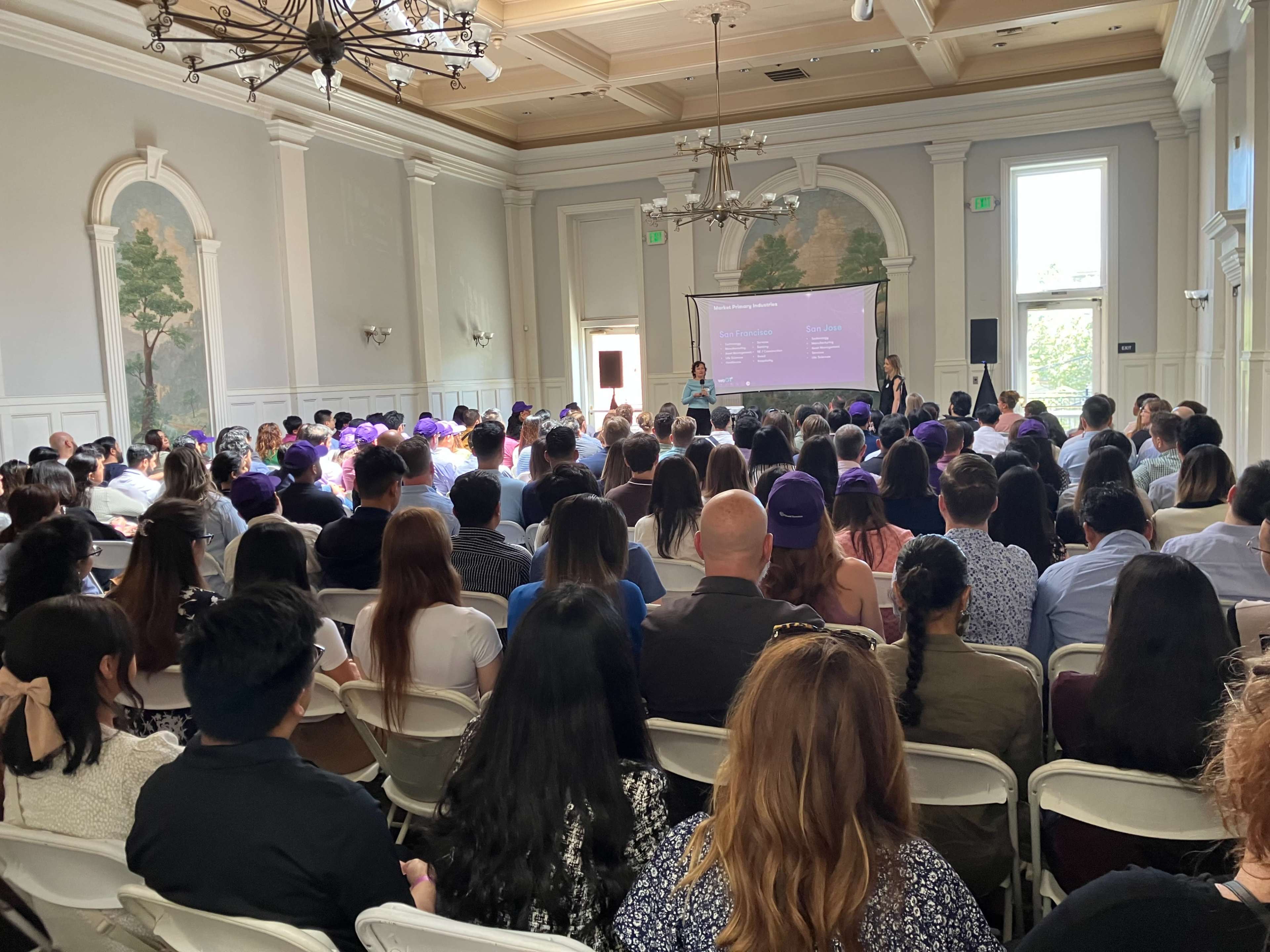 A large audience is seated in a conference room, facing a presentation screen, with two speakers at the front.