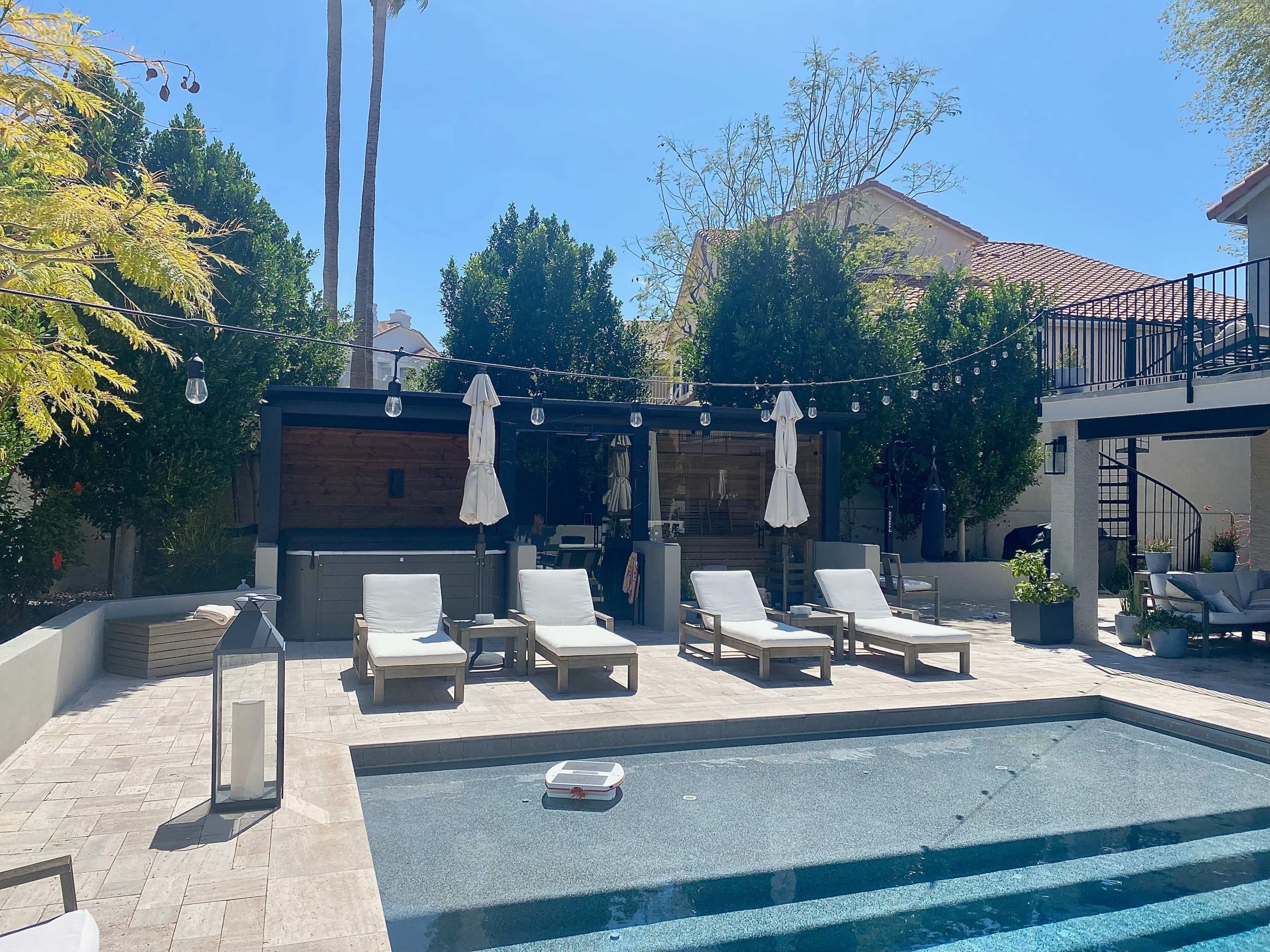 A modern backyard pool area with lounge chairs, a pool, and a covered outdoor seating space, all surrounded by landscaped greenery and trees.