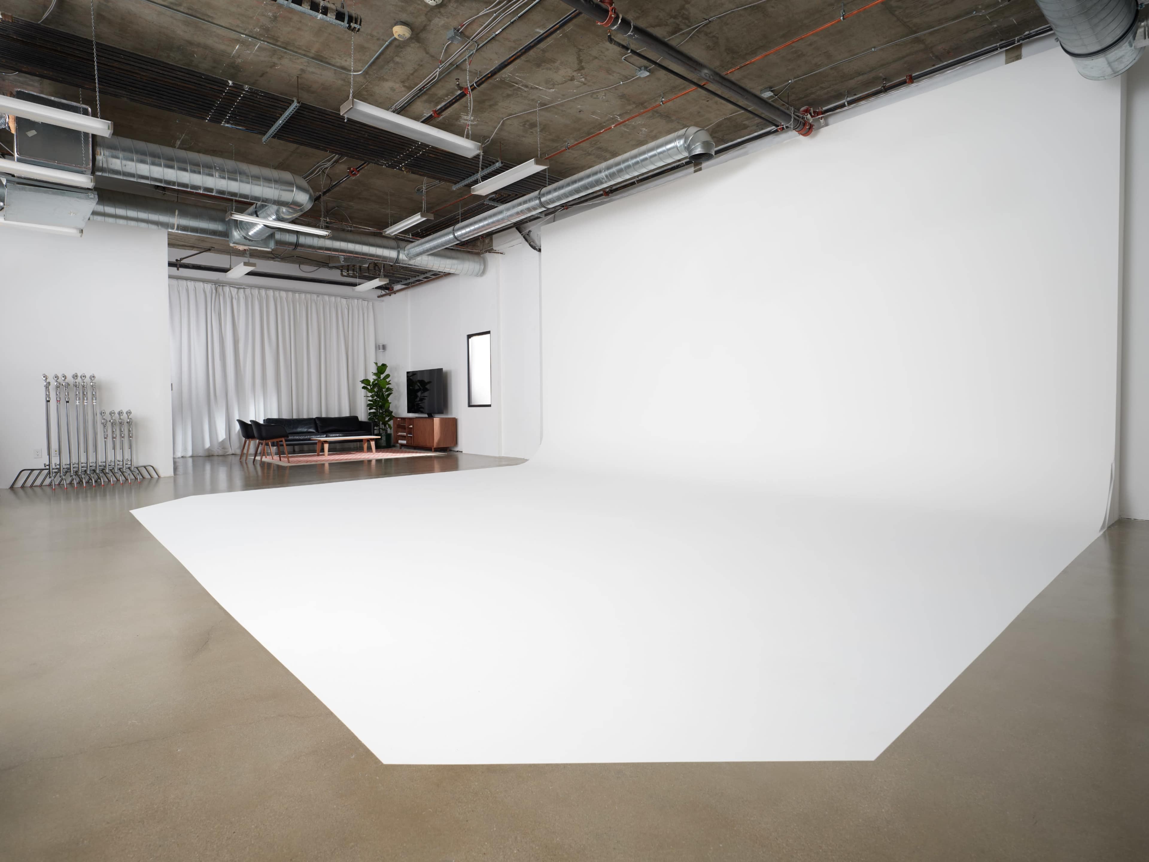 A large, minimalistic studio space with a seamless white backdrop, polished concrete floors, and a black sofa in one corner.