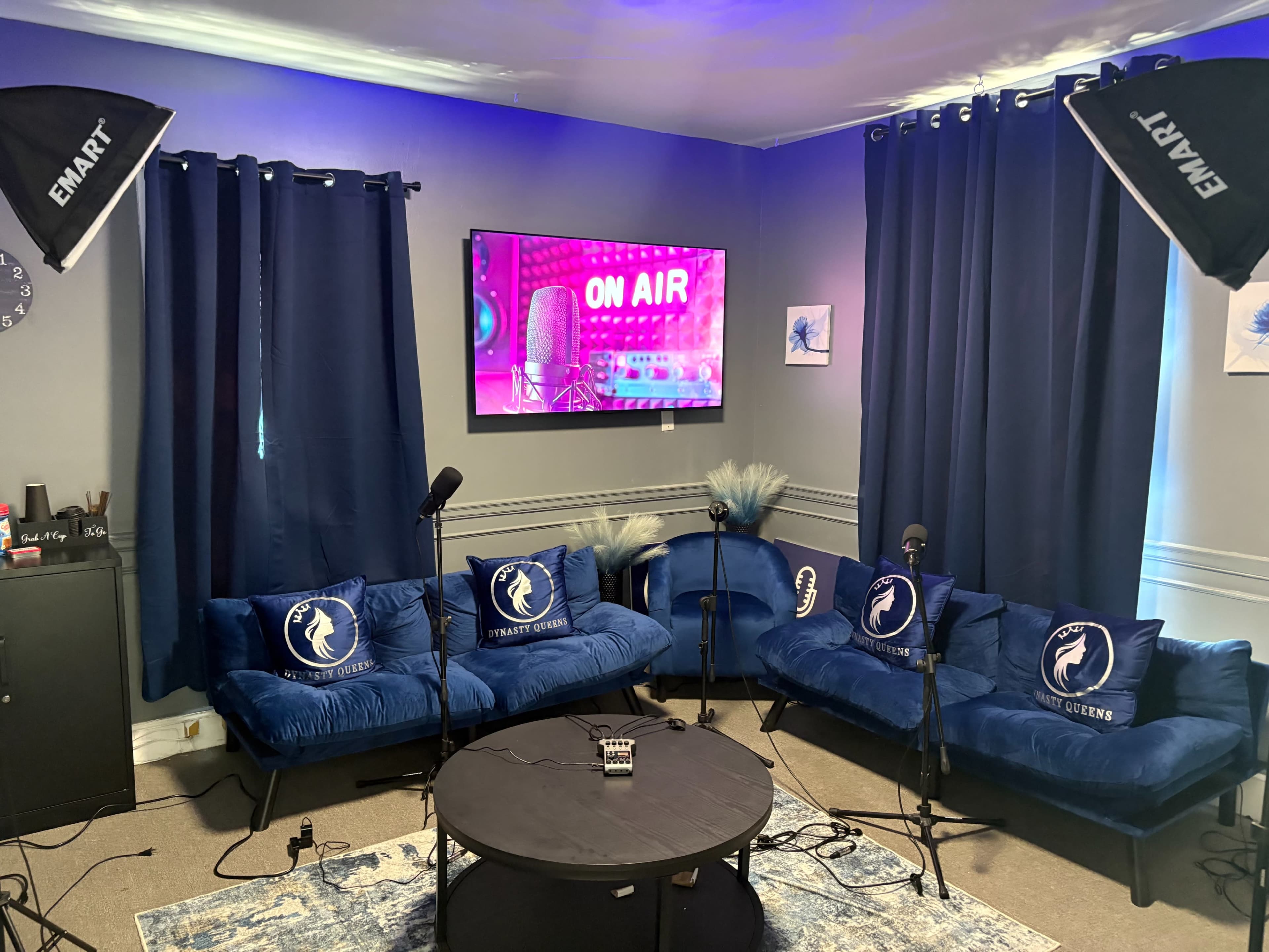 The image shows a cozy studio setup with a blue velvet sectional couch, two microphones, and a large TV displaying "ON AIR," all surrounded by dark curtains and studio lights.