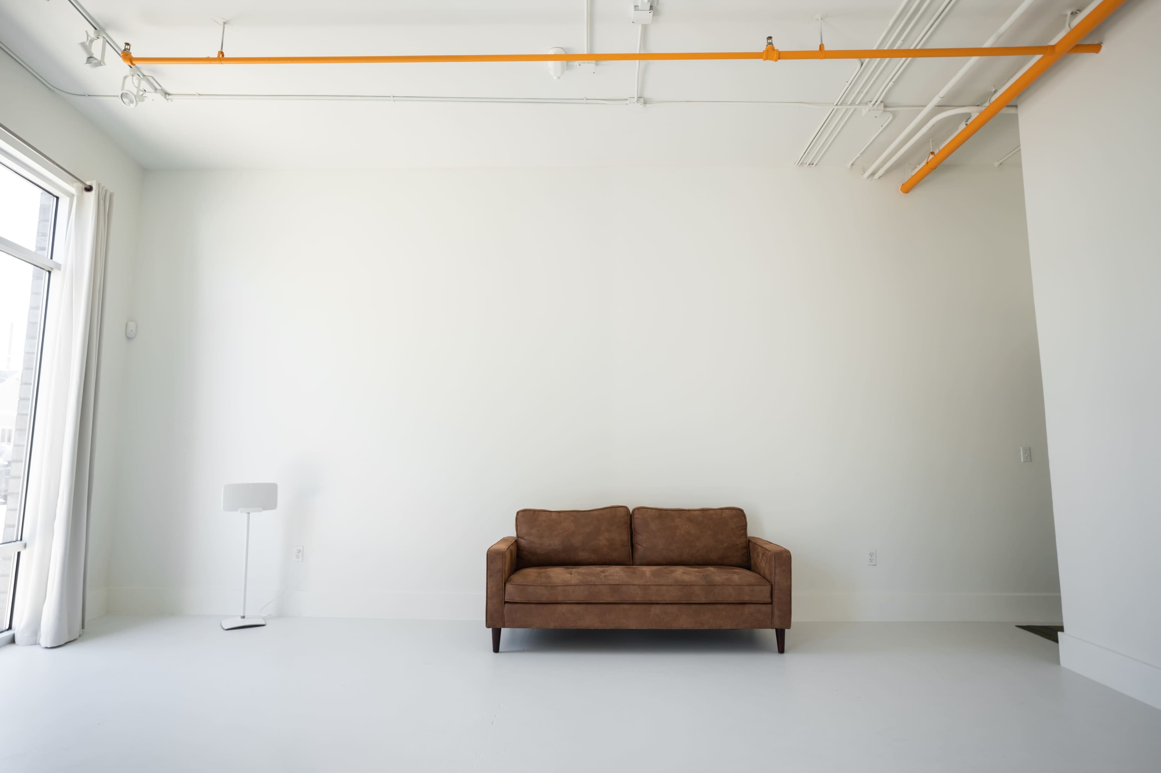 A minimalist, brightly lit interior with a brown sofa centered against a blank white wall, accompanied by a floor lamp in the corner.