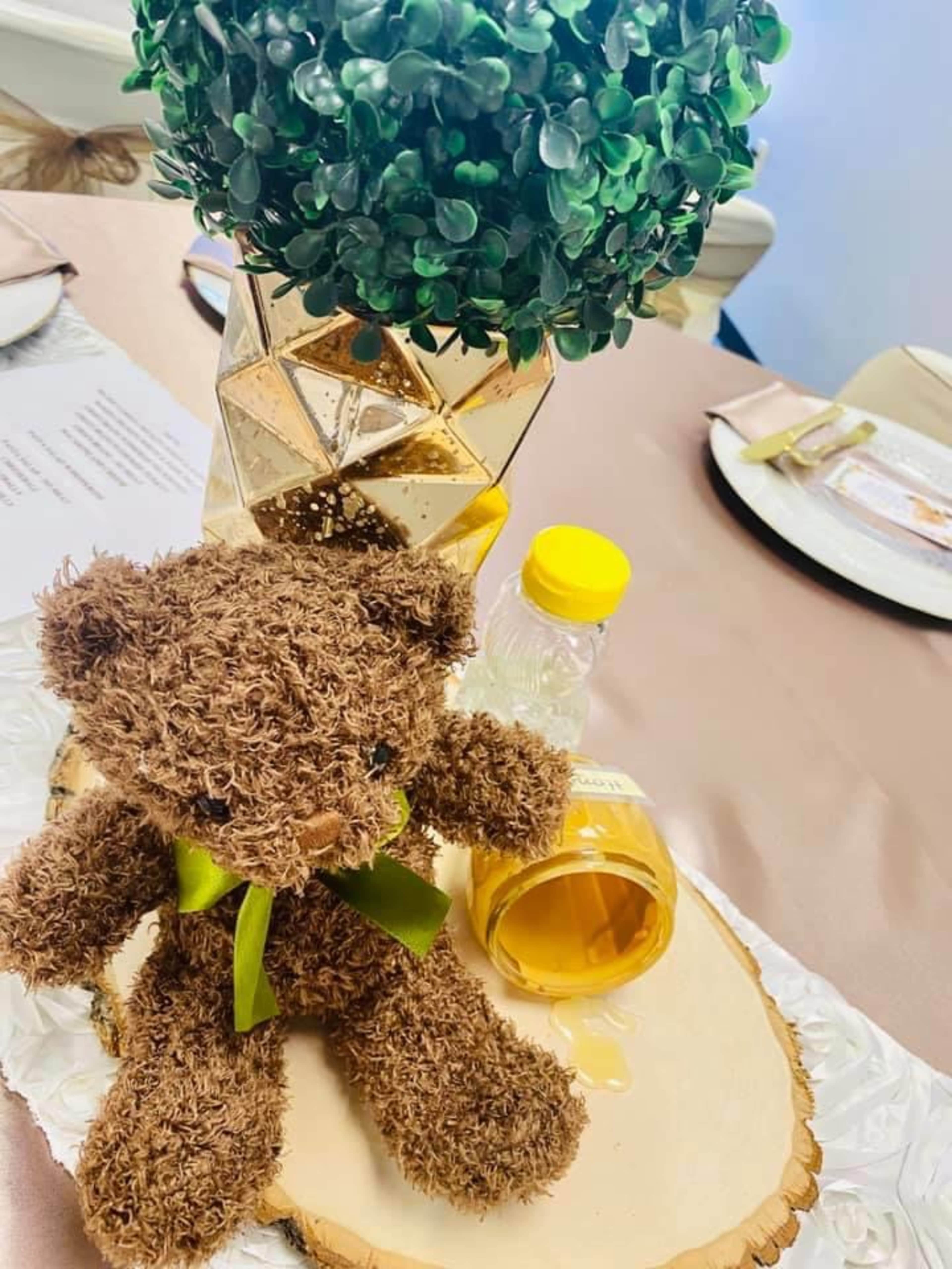 A brown teddy bear sits next to a bottle of oil on a wooden platter, with a decorative plant in a geometric vase in the background.