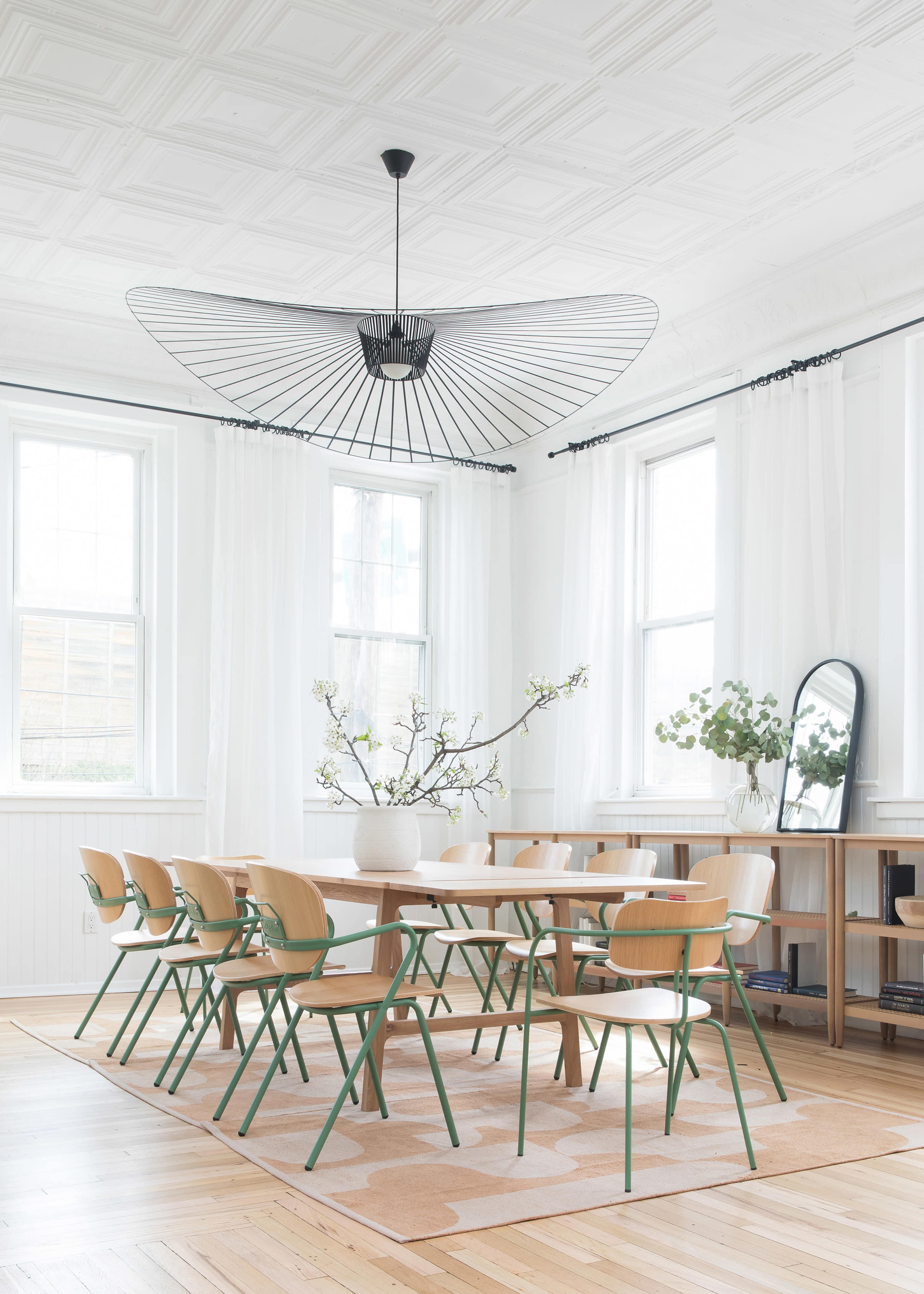 A dining room features a wooden table surrounded by eight chairs, with large windows adorned by sheer curtains and a decorative pendant light overhead.
