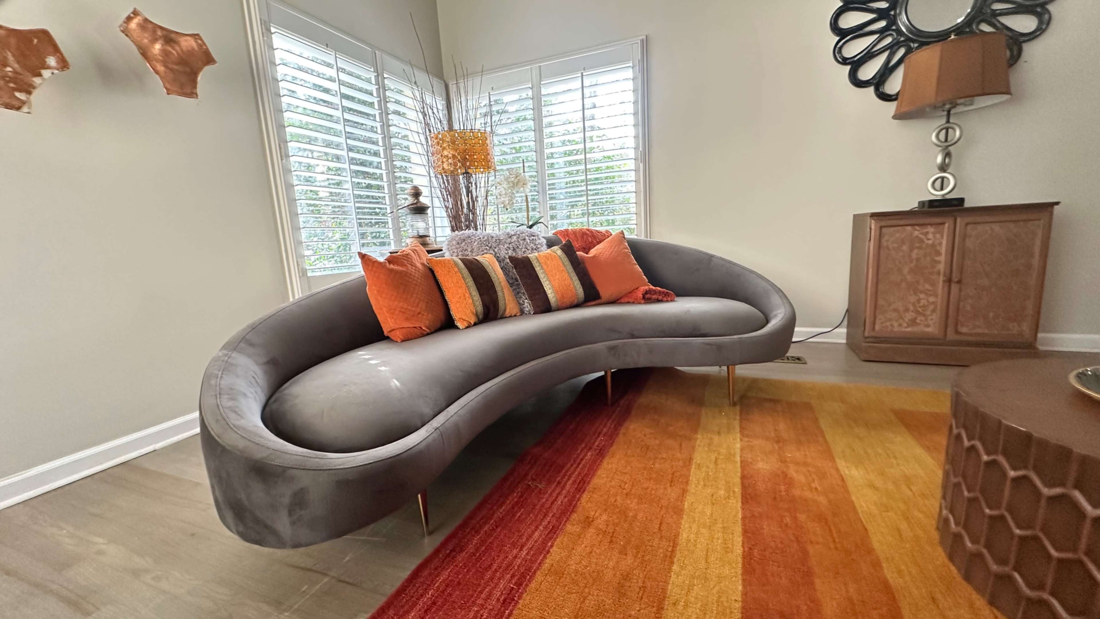 The image shows a modern, curved gray sofa adorned with orange and brown pillows, set against a backdrop of window shutters and a patterned area rug.