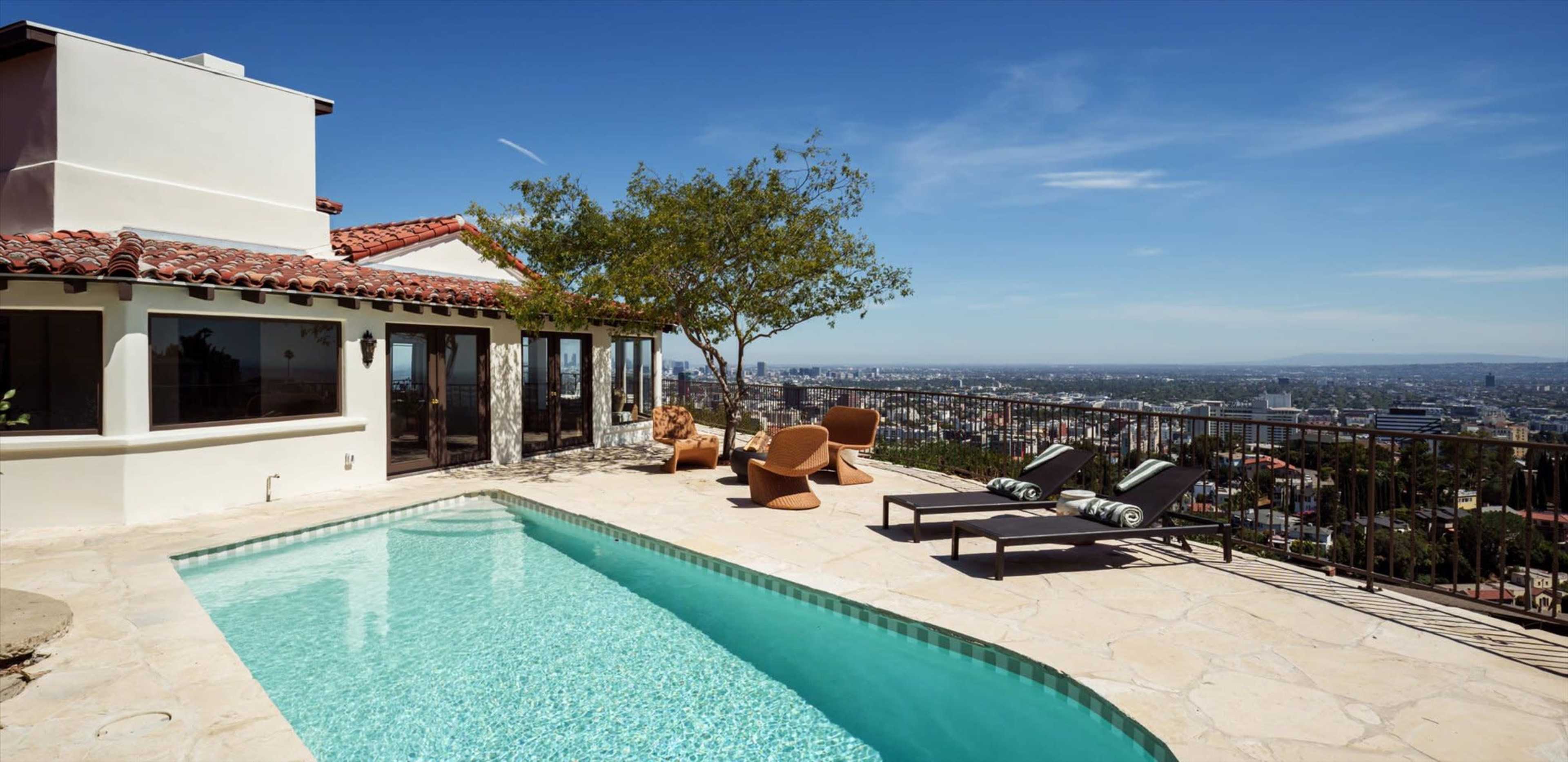 The image shows a poolside view of a modern villa overlooking a cityscape, with lounge chairs and a tree in the background under a clear blue sky.