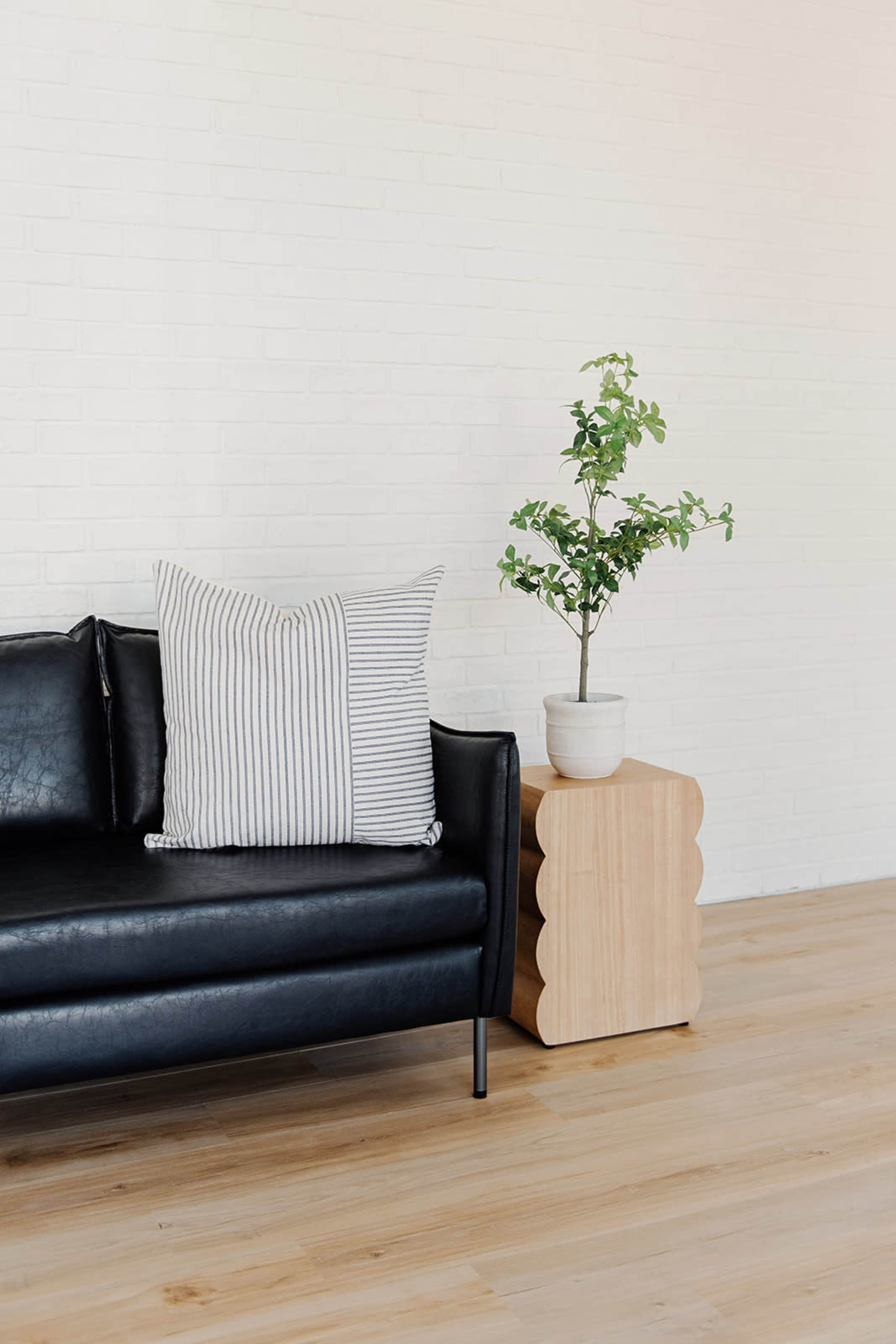A black sofa with striped pillows is positioned next to a wooden side table that holds a potted plant against a white brick wall.