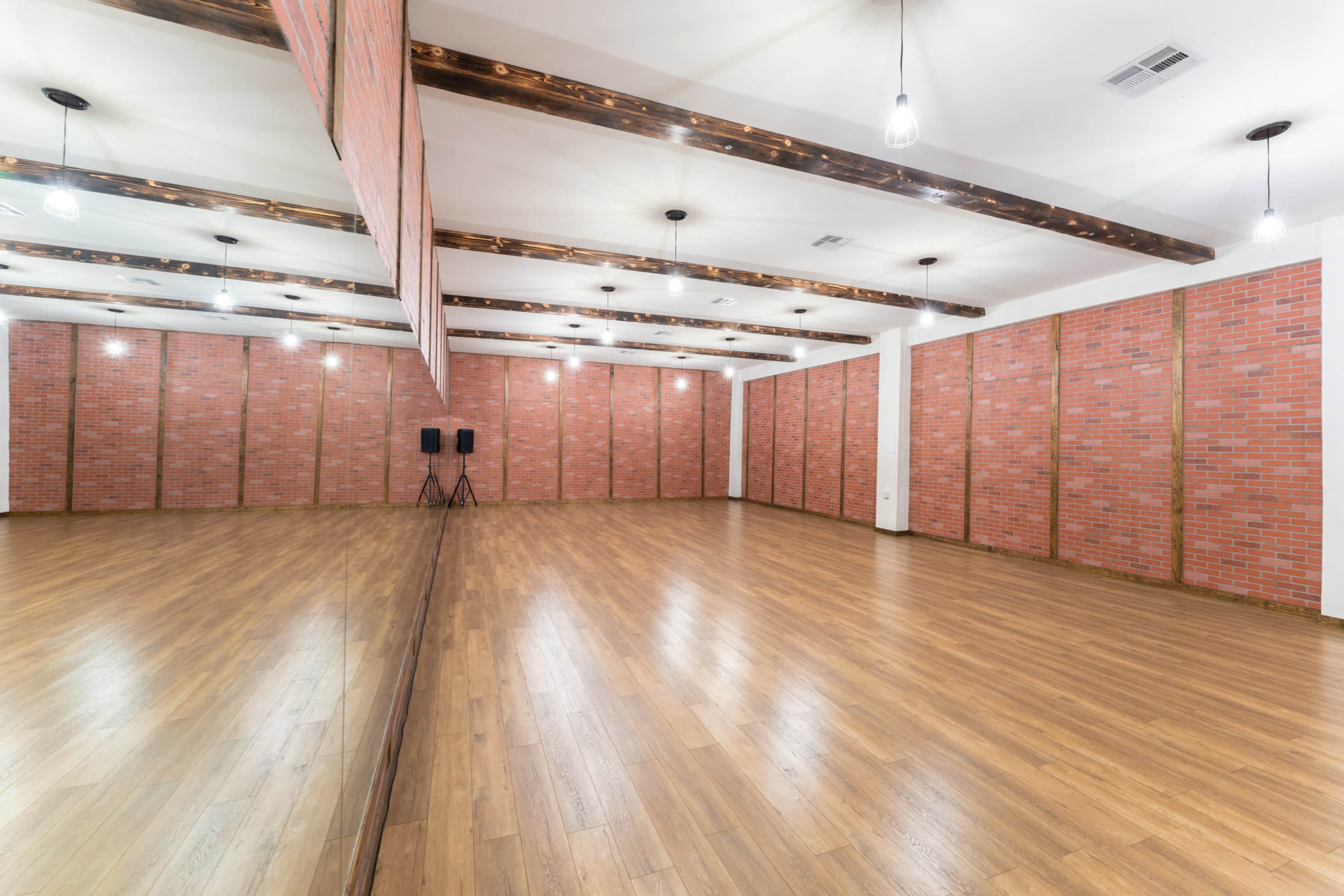 The image shows a spacious, empty room with wooden floors and exposed brick walls, featuring ceiling lights and a large mirror along one side.
