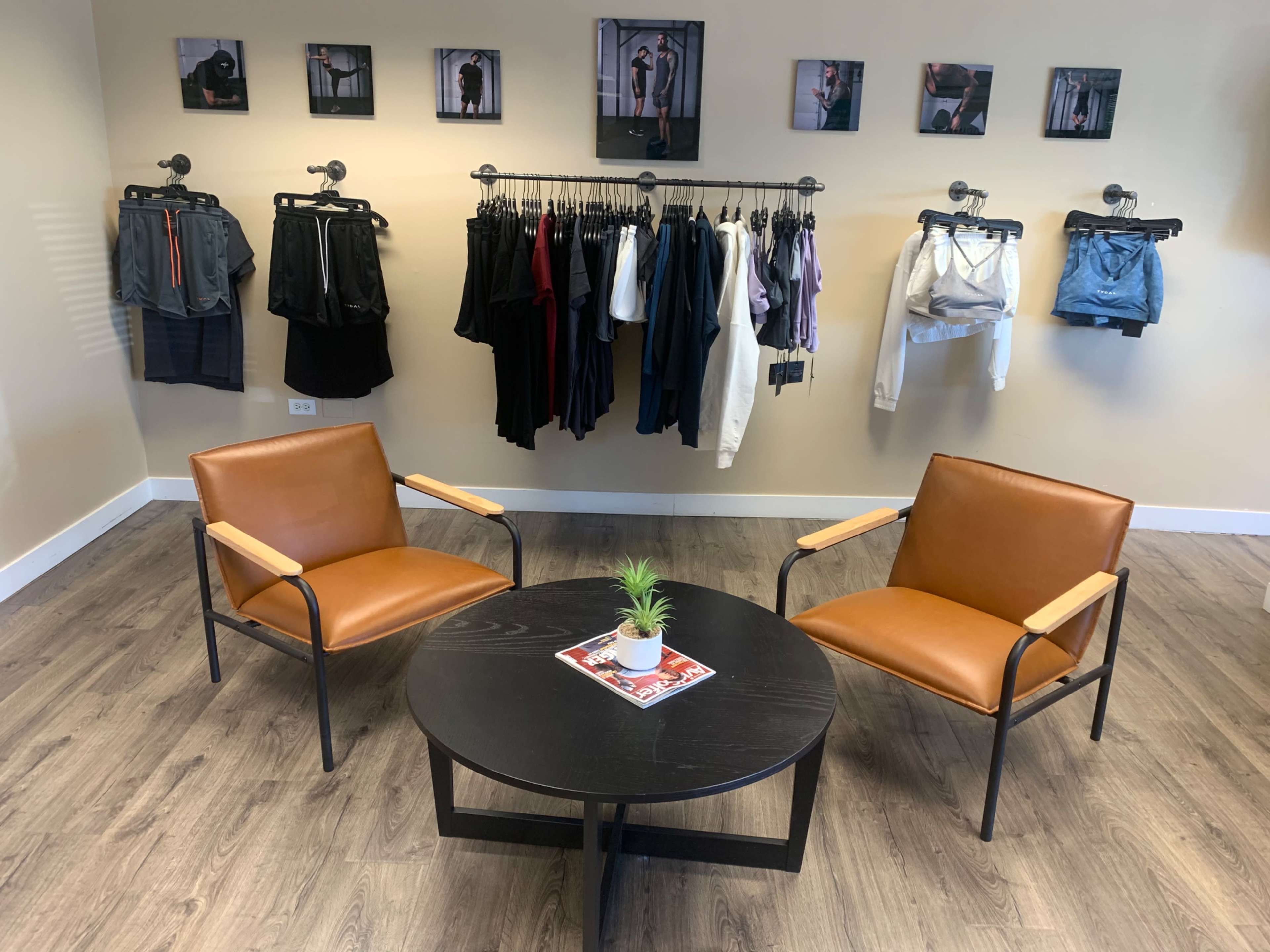 A well-lit room with two brown chairs and a black table in the center, surrounded by clothing displayed on hangers along the wall.