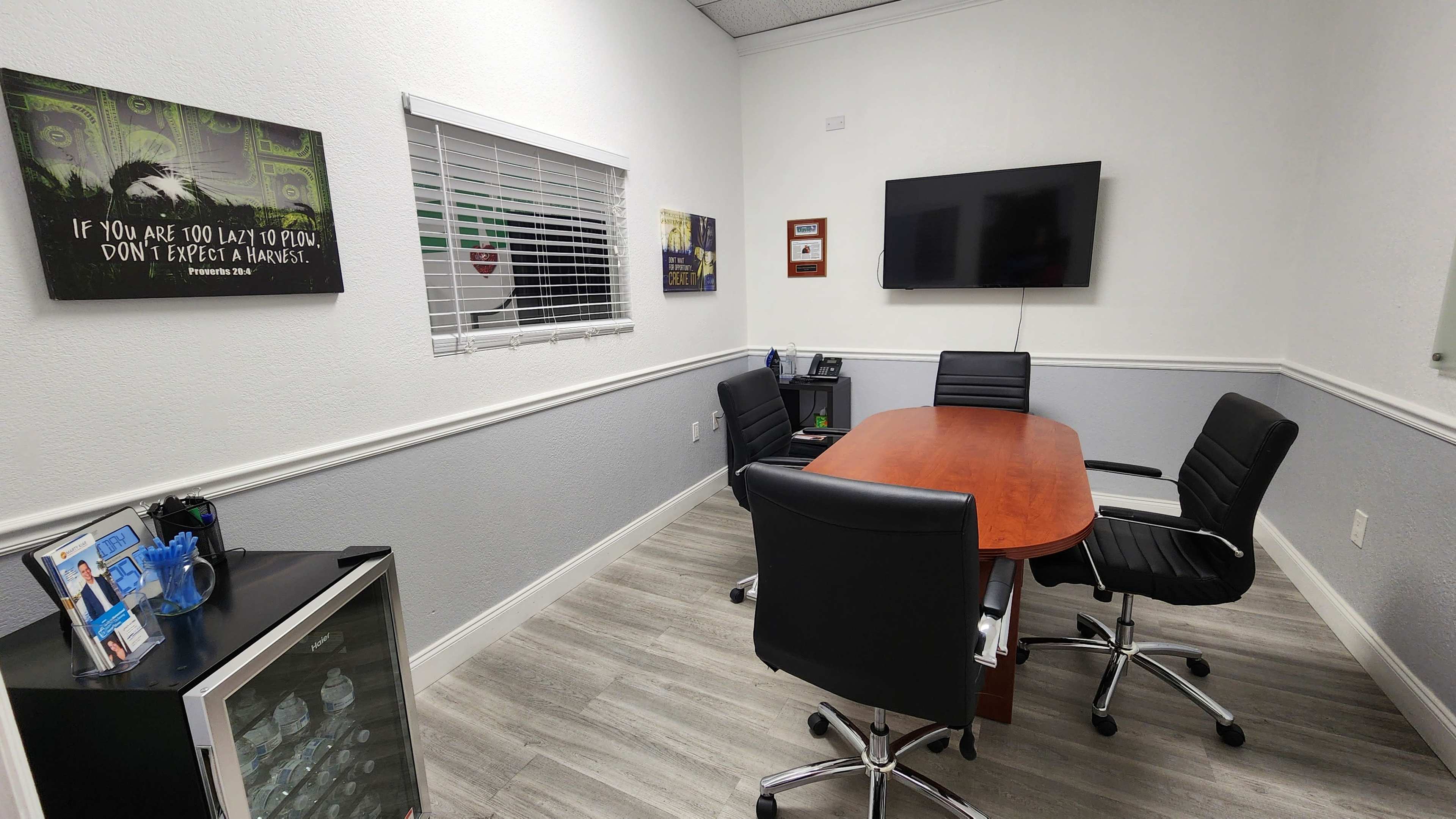 A small conference room with a wooden table surrounded by black office chairs, a television mounted on the wall, and various framed posters.