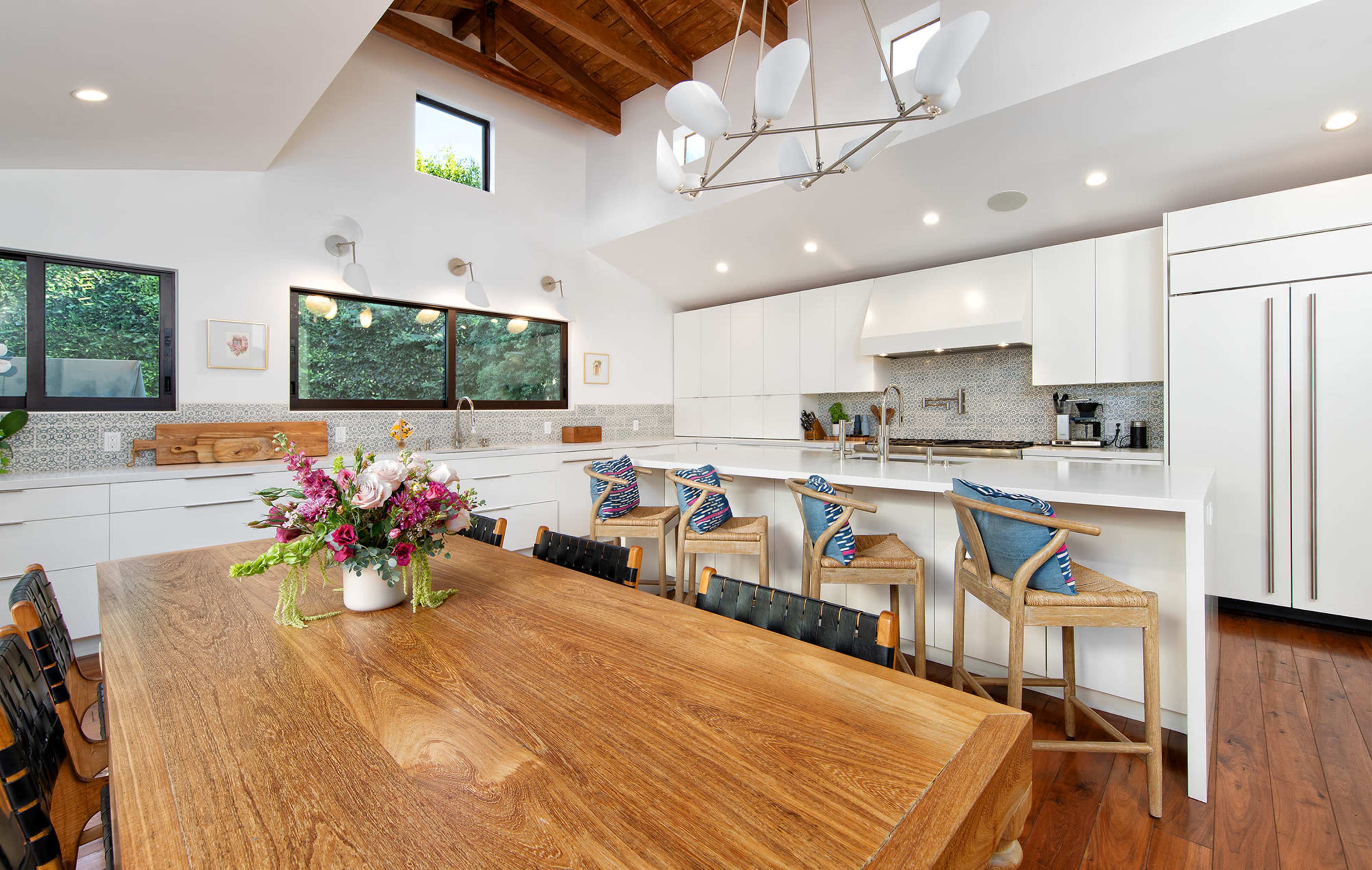 A bright, modern kitchen features white cabinetry, a large wooden dining table, and barstools with colorful backs, illuminated by natural light from large windows.
