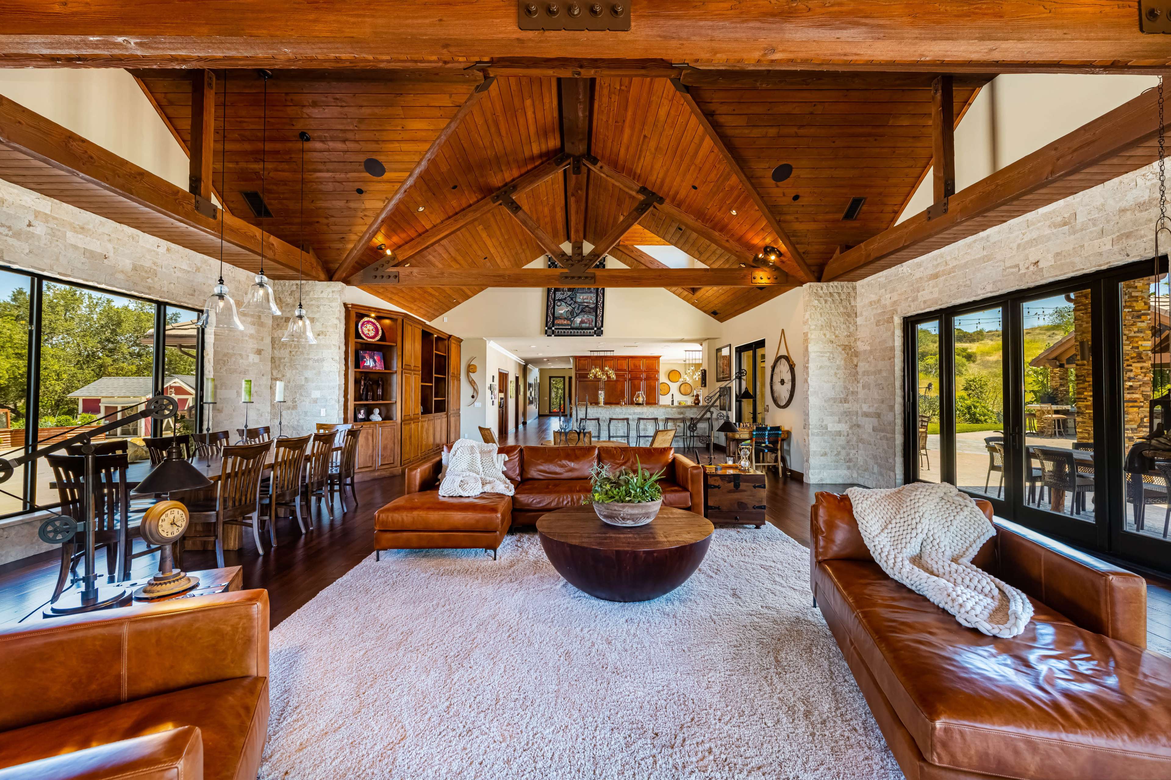The image shows a spacious living room with exposed wooden beams, large windows, leather sofas, and a round coffee table on a light rug.