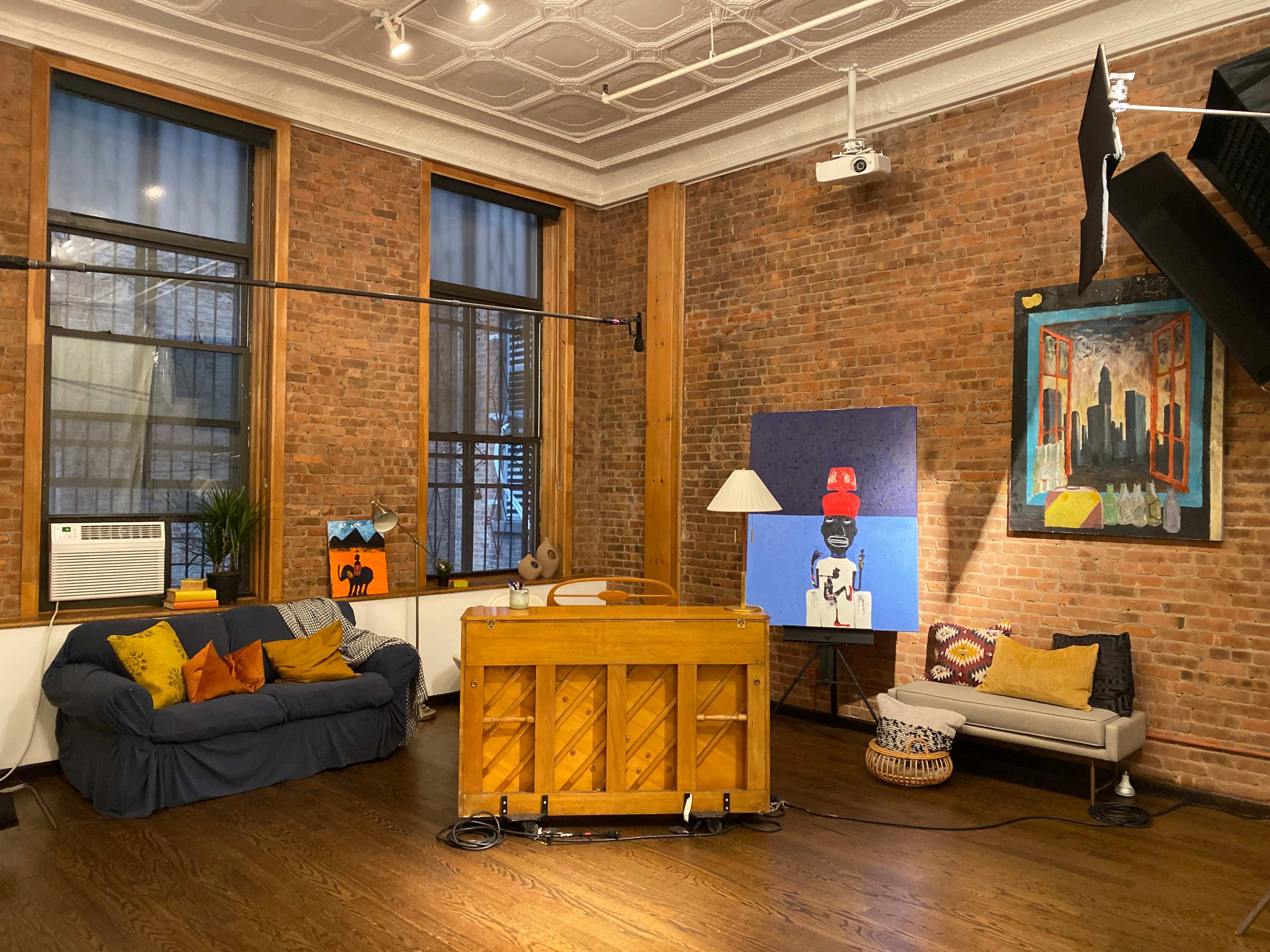 The image shows a cozy room with exposed brick walls, two windows, a piano, and various artworks on display.
