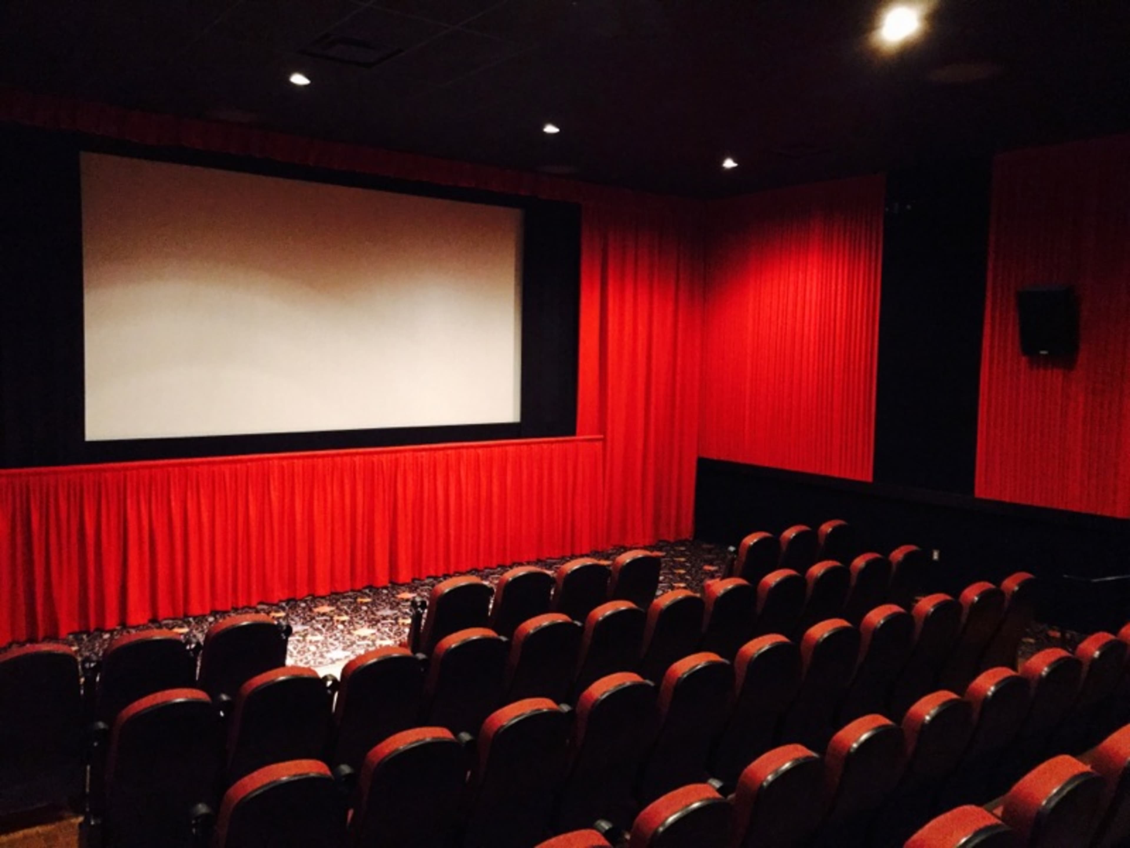 An empty movie theater with red curtains, a large screen, and rows of cushioned seats.