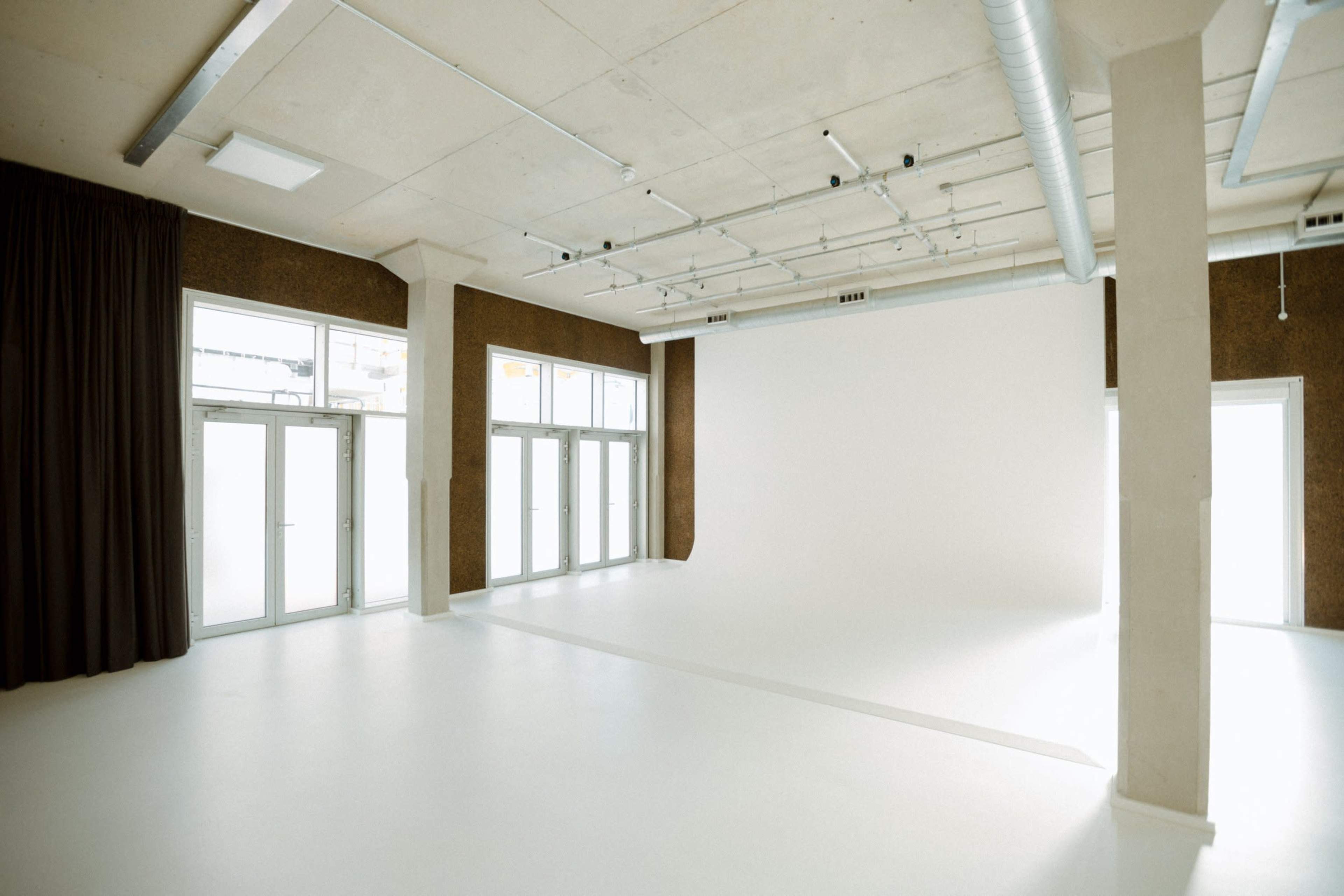 A spacious studio with large windows, white walls, and a blank backdrop, featuring a polished floor and exposed ceiling infrastructure.