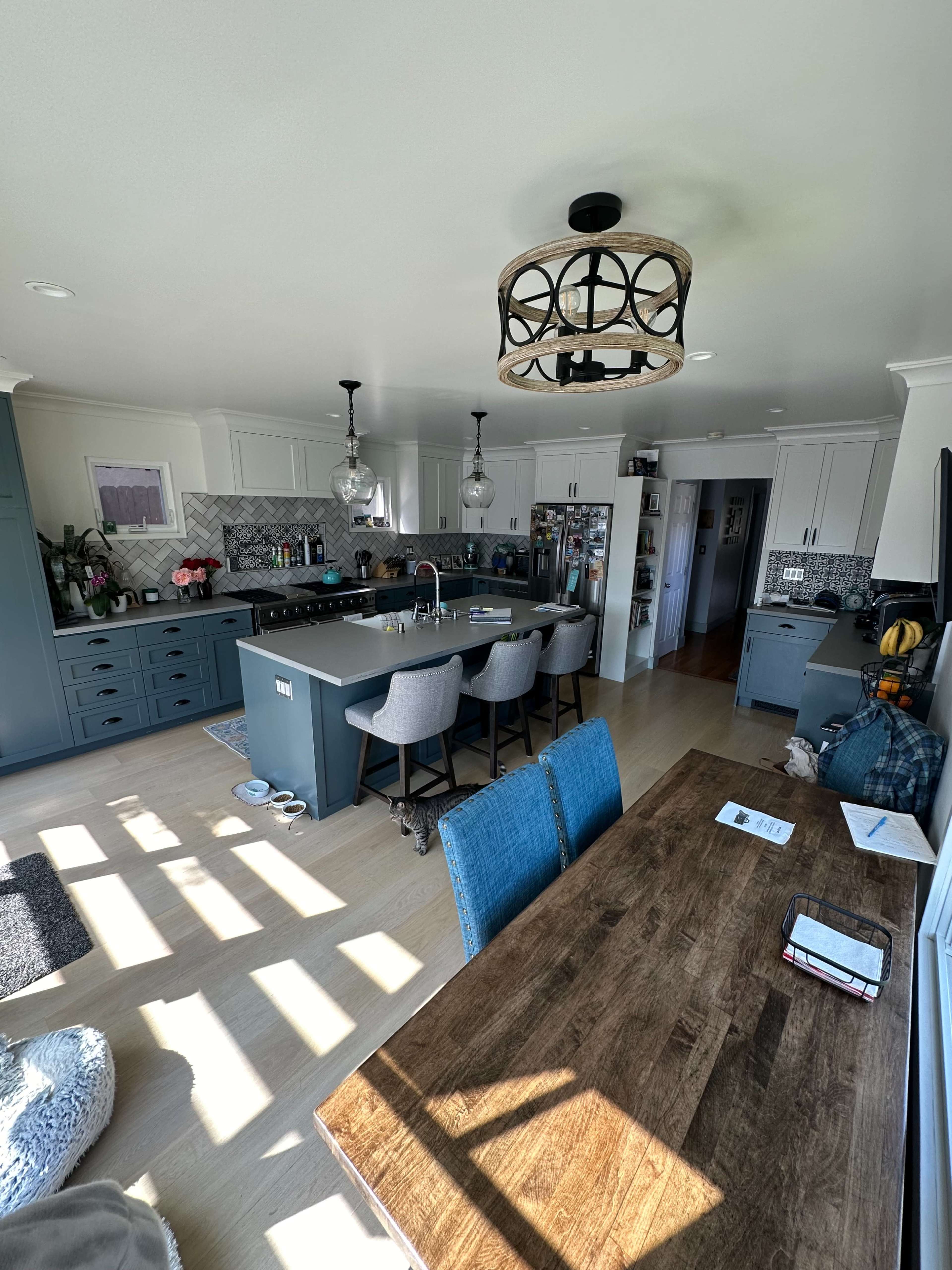The image shows a modern kitchen with a large island, bar stools, and a dining table, all illuminated by natural light streaming through windows.