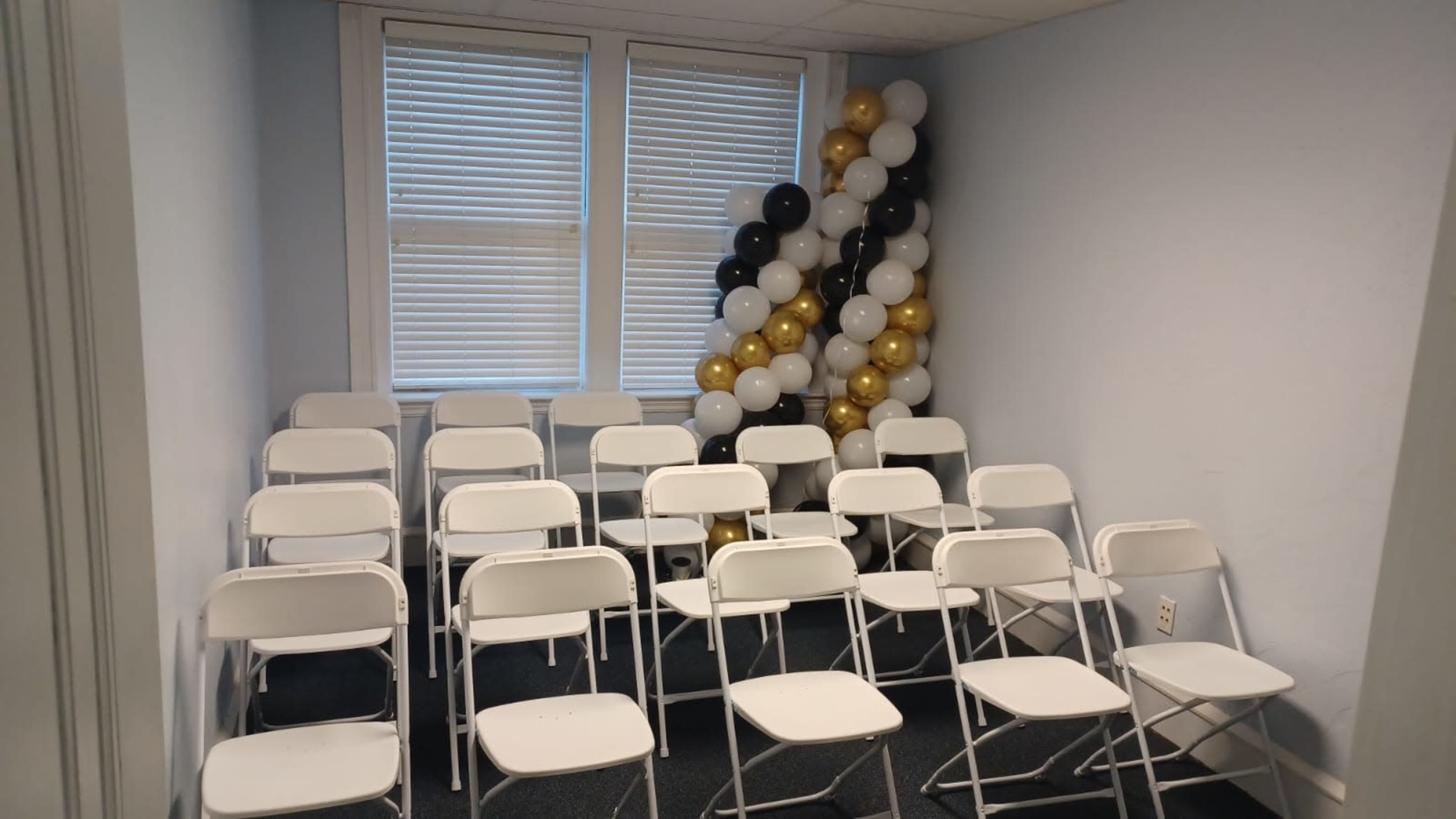 A small room is set up with rows of white folding chairs facing a cluster of black, white, and gold balloons.