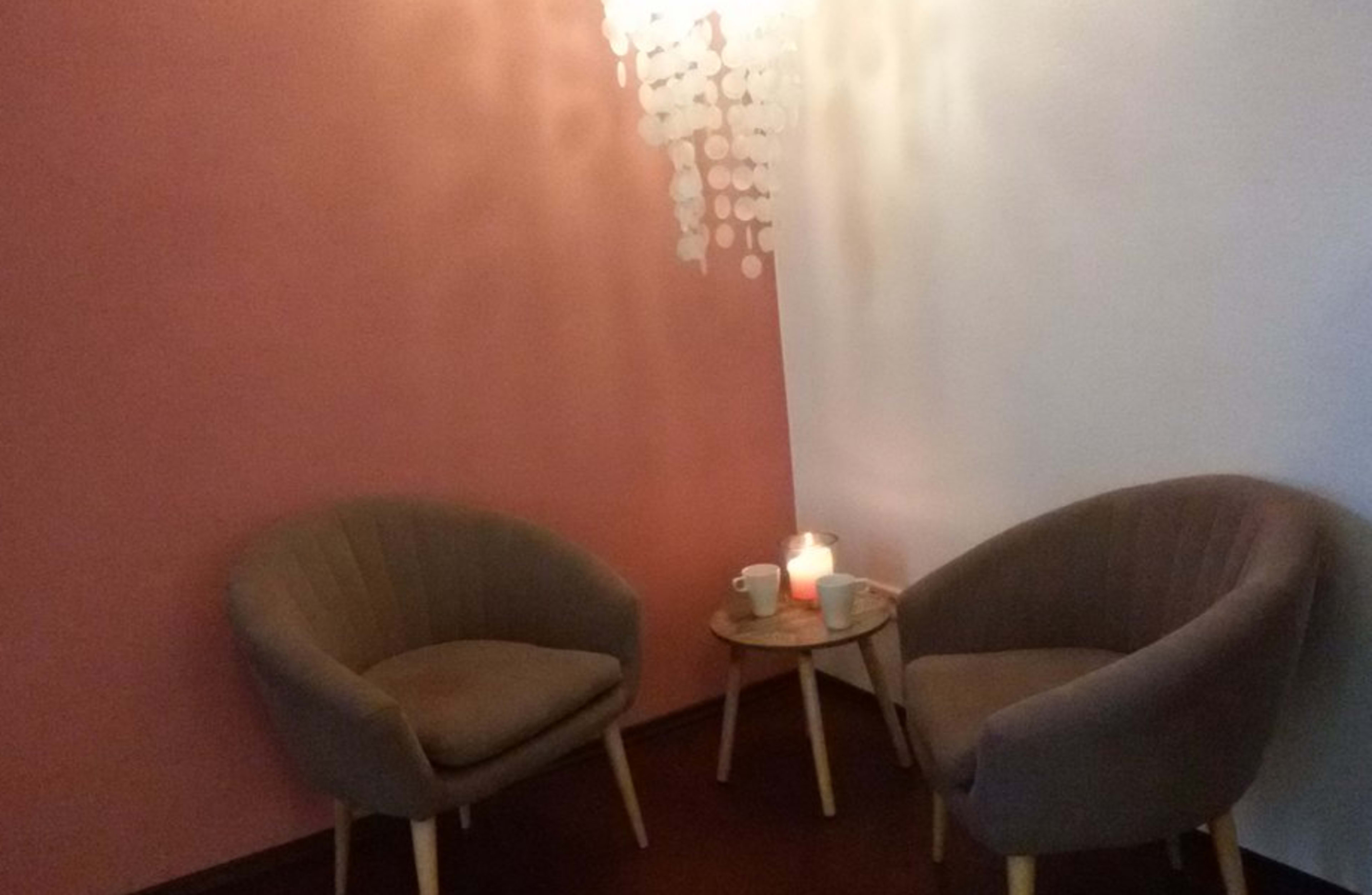 A cozy corner with two upholstered chairs, a small circular table holding a candle and a cup, and a decorative hanging element in the background.
