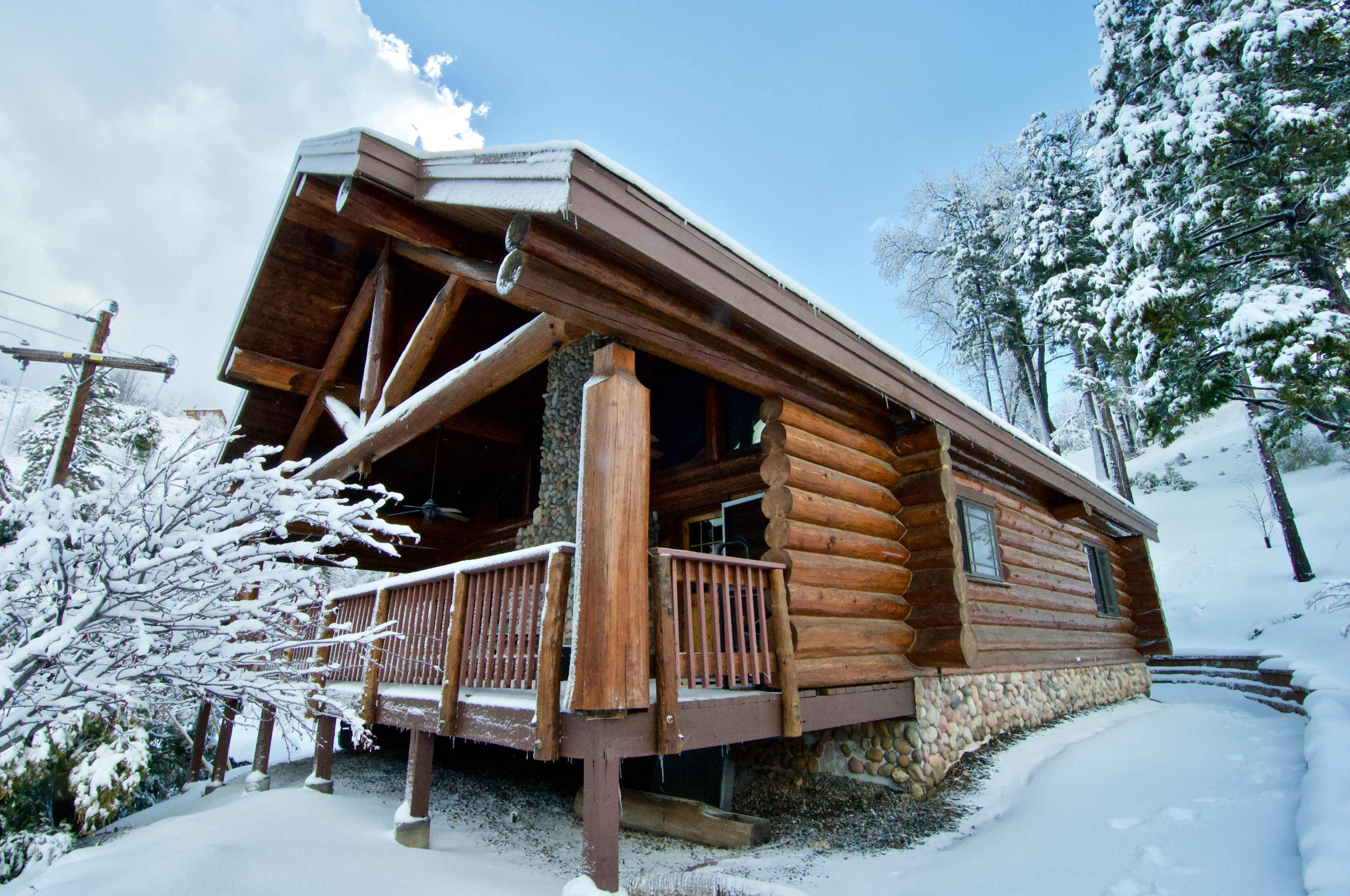 A log cabin with a stone foundation is surrounded by snow-covered trees under a partly cloudy sky.