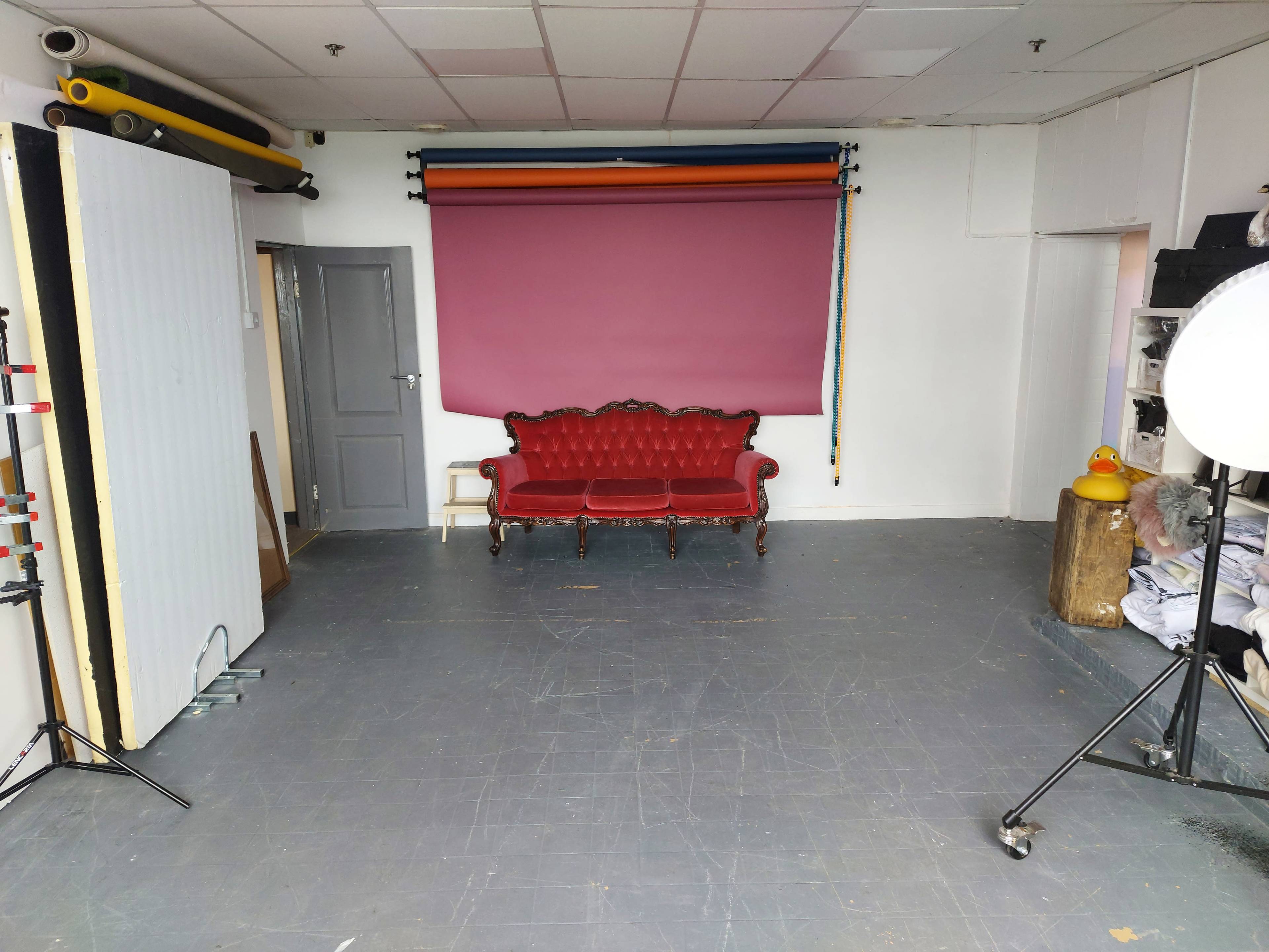 A red vintage couch is centered in a spacious studio with a pink backdrop and various photography equipment nearby.