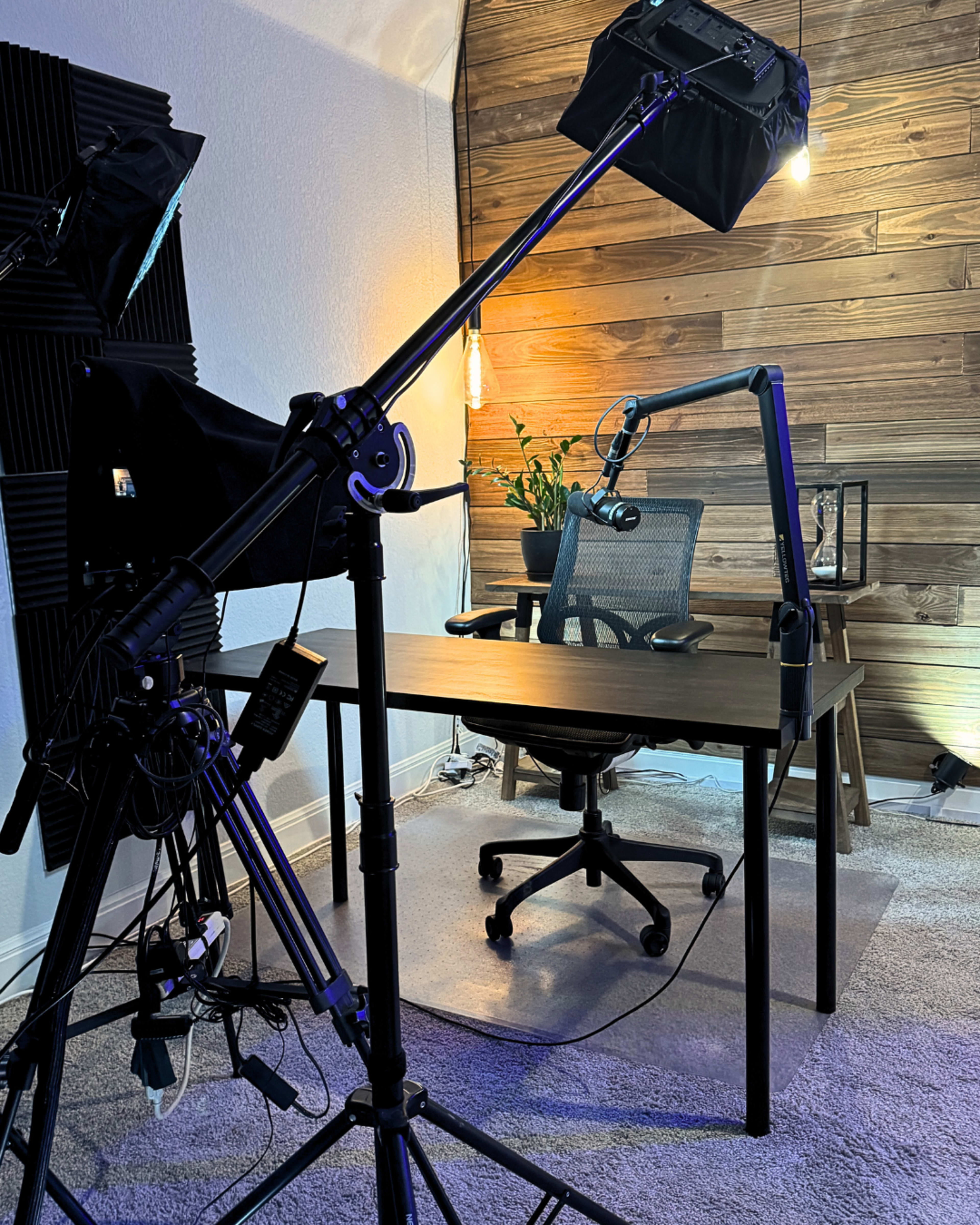 The image shows a well-lit office setup with a desk, an ergonomic chair, and camera equipment positioned to capture the scene.