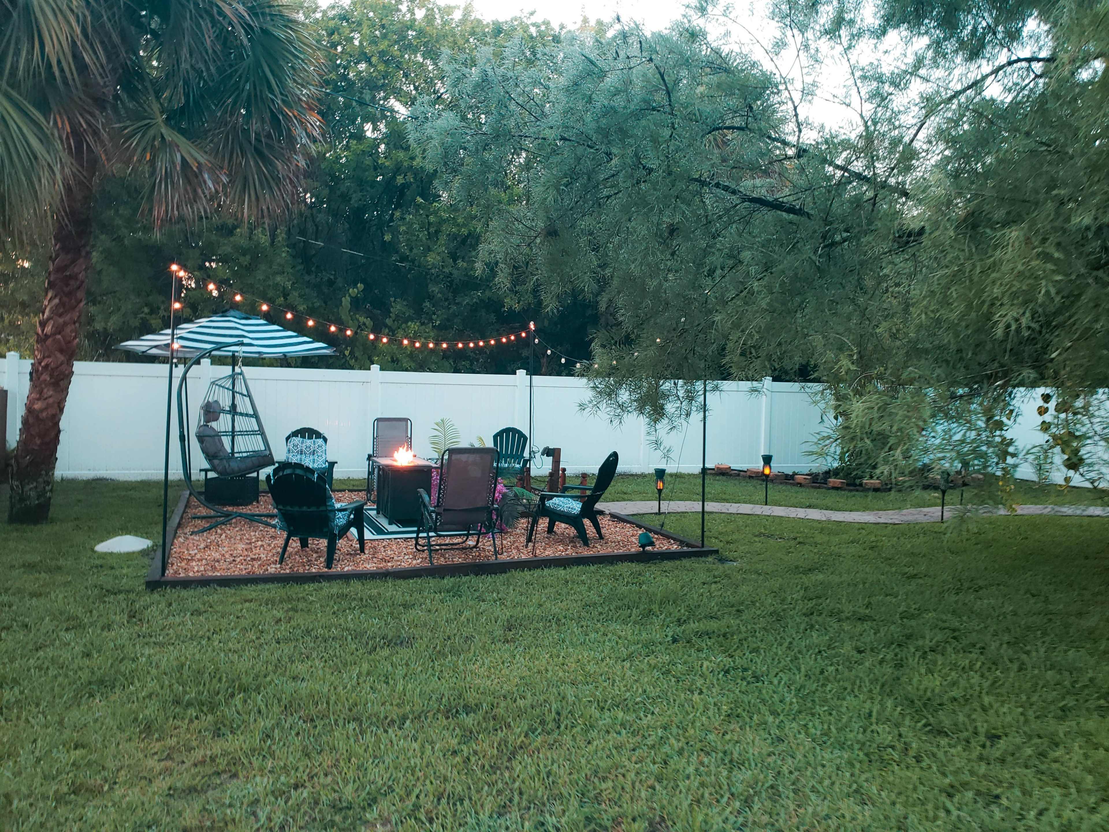 A backyard patio area features chairs arranged around a fire pit, string lights, and a shaded sitting area.