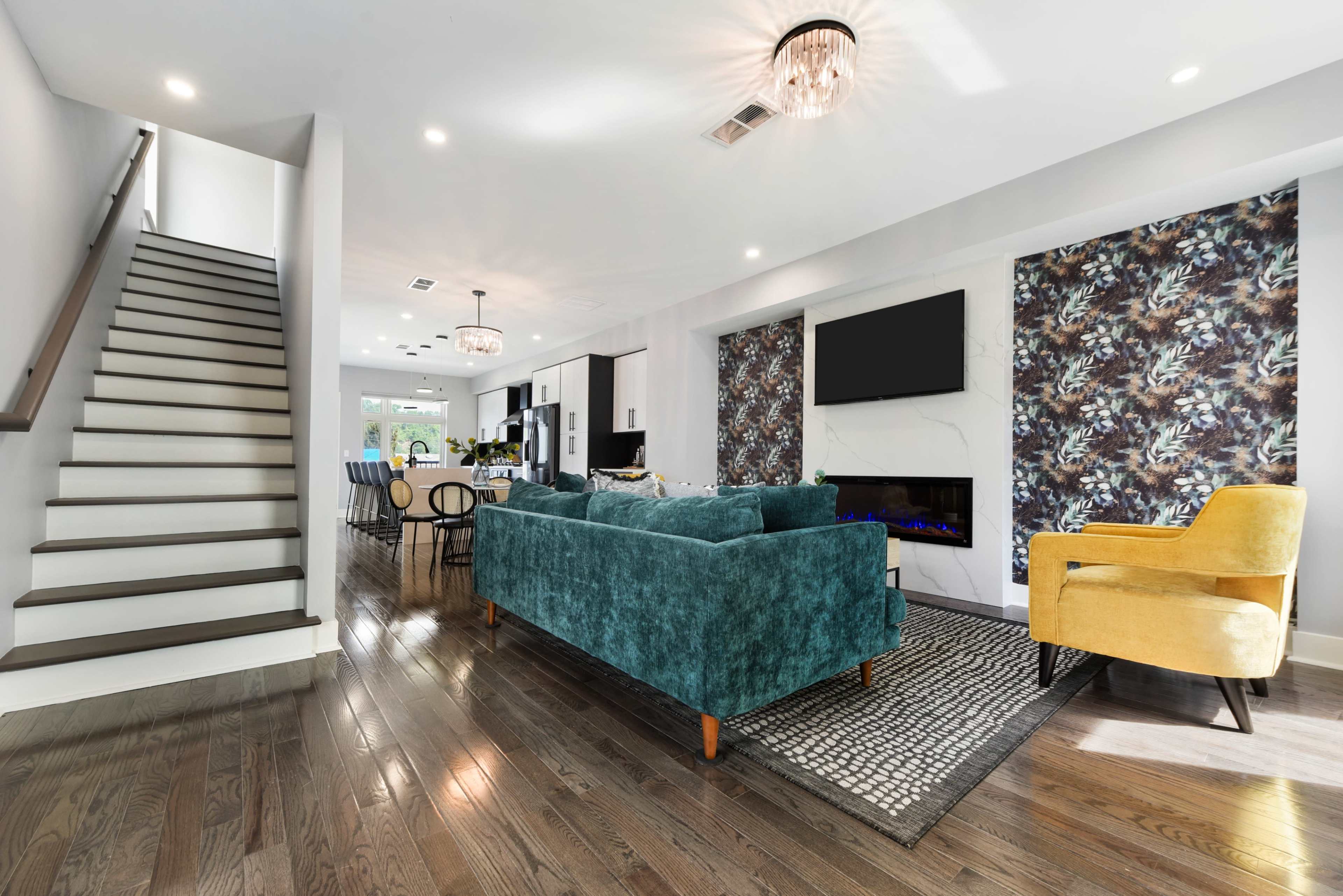 A modern living room features a teal sofa, yellow armchair, and a staircase leading to an upper level, with a fireplace and a patterned accent wall.