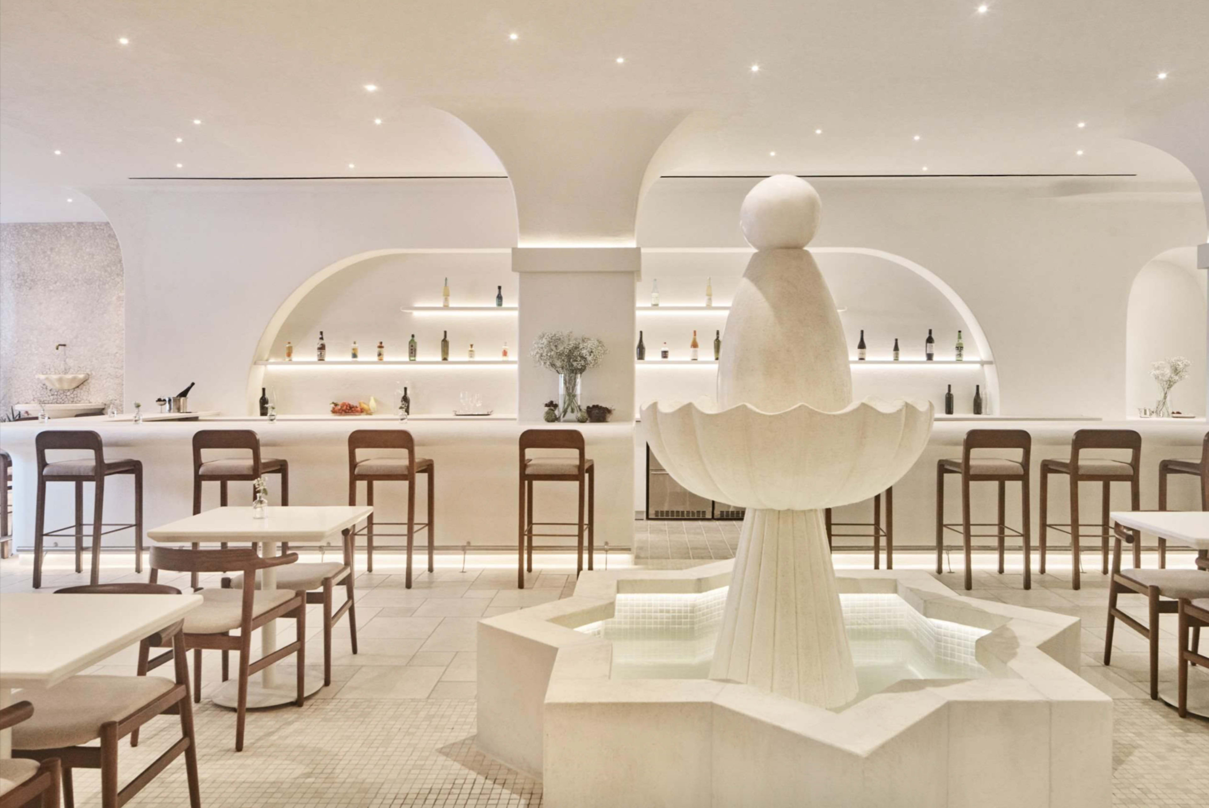 The image shows a modern restaurant interior featuring a central decorative fountain, a minimalist bar lined with bottles, and several light-colored tables and chairs.