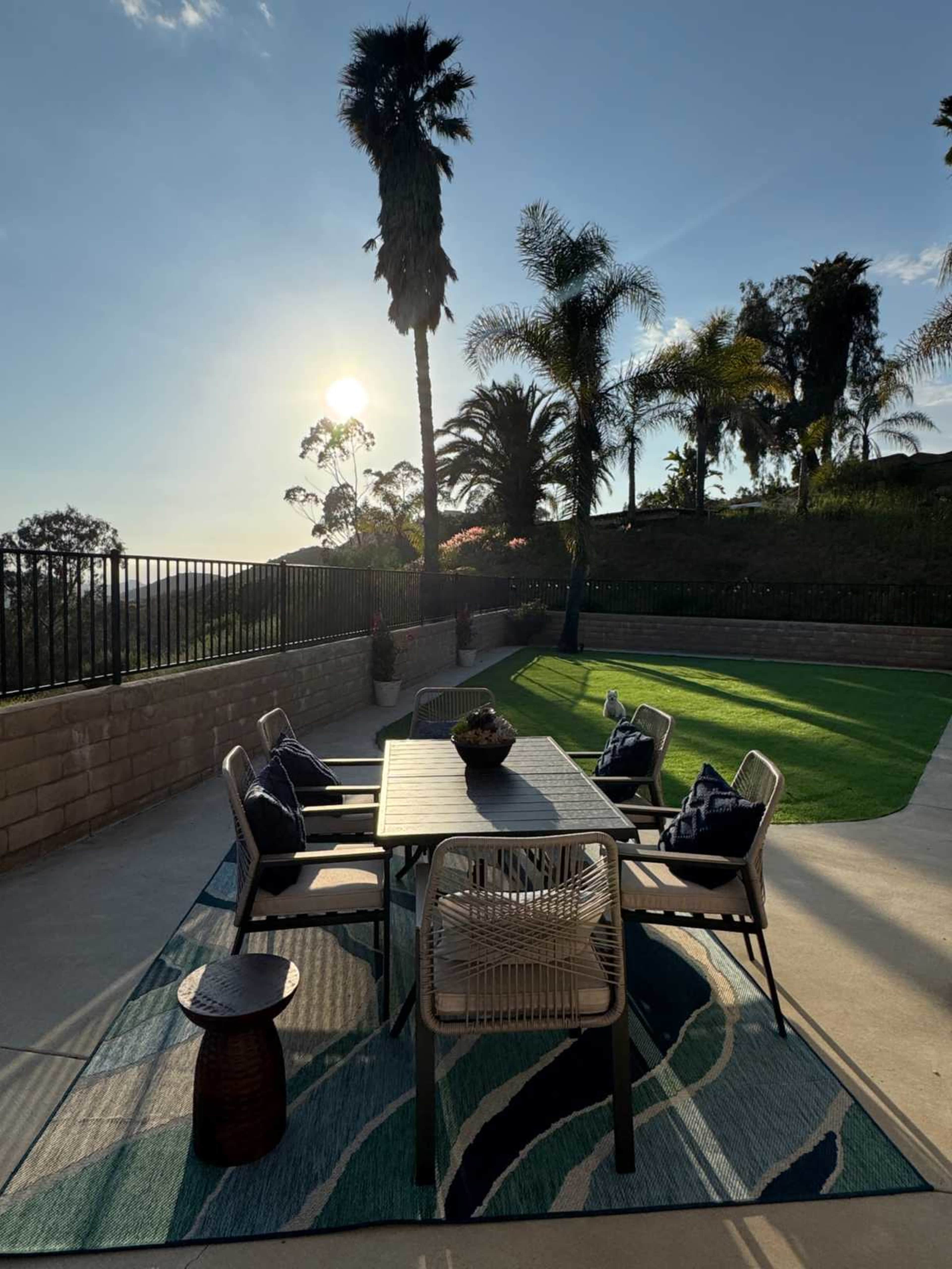 A dining table with chairs is set on a patio next to a lawn, with palm trees and a sunset in the background.